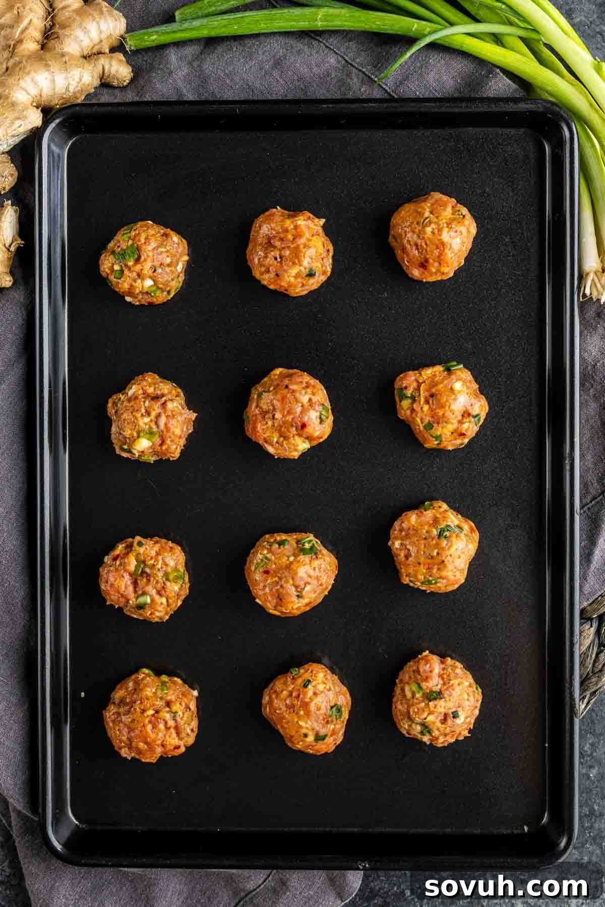 Effortless Viral Blanket Dumplings No Folding Needed 5 Twelve uncooked pork dumplings with herbs are evenly spaced on a black baking sheet, with fresh ginger and green onions visible in the background—perfect for making no fold dumplings or wonton wrapper dumplings.