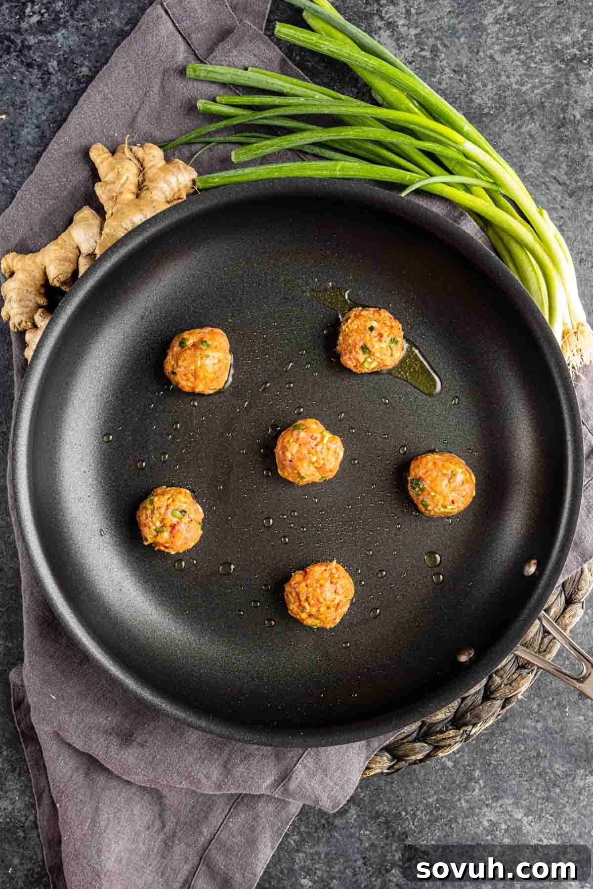 Effortless Viral Blanket Dumplings No Folding Needed 6 Six uncooked meatballs in a black skillet with a bit of oil, placed on a dark cloth next to fresh ginger and green onions—perfect for making viral blanket dumplings or wonton wrapper dumplings on a dark textured surface.
