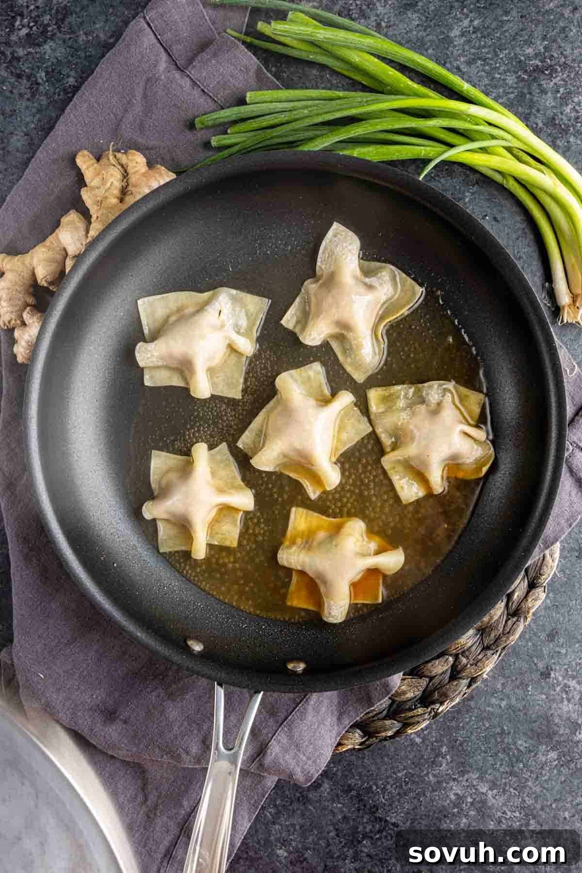 Effortless Viral Blanket Dumplings No Folding Needed 7 Six pork dumplings are being pan-fried in oil in a black skillet; ginger root and green onions are placed nearby on a dark countertop.