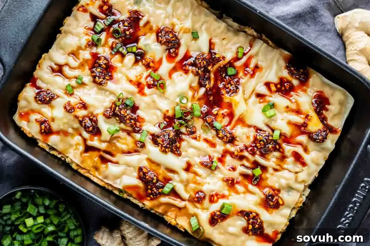 Viral Wonton Lasagna Twist 2 A baked viral dumpling lasagna in a black rectangular pan, topped with chili oil, sauce, and chopped green onions, with extra green onions in a bowl on the side.