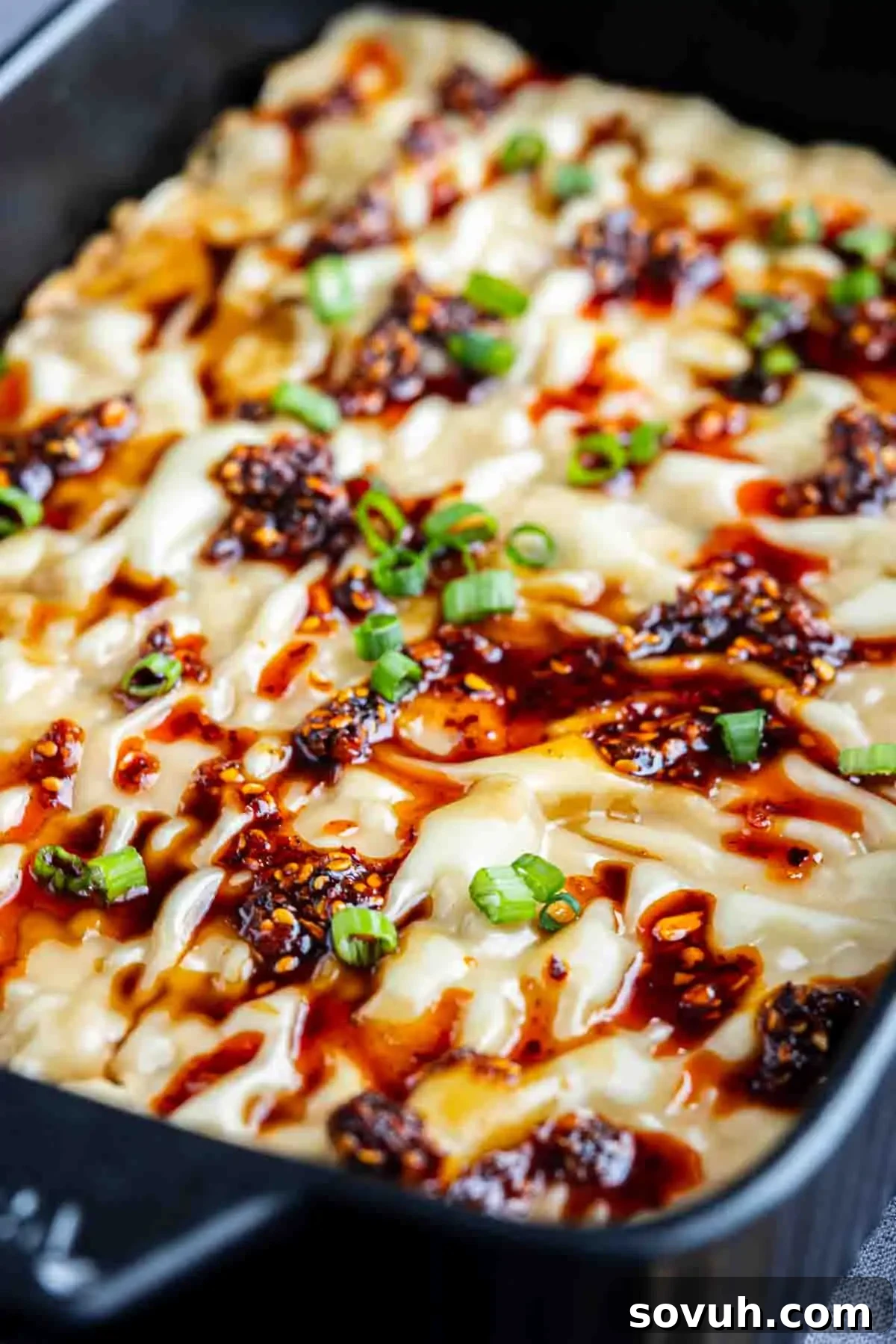 Viral Wonton Lasagna Twist 11 A close-up of a baked TikTok dumpling lasagna topped with melted cheese, chili oil, and chopped green onions in a black baking dish.