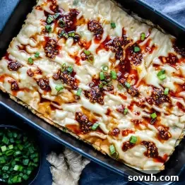 Viral Wonton Lasagna Twist 12 Rectangular baking dish with layered viral dumpling lasagna topped with spicy chili oil and chopped green onions, next to a bowl of extra green onions and a piece of ginger.