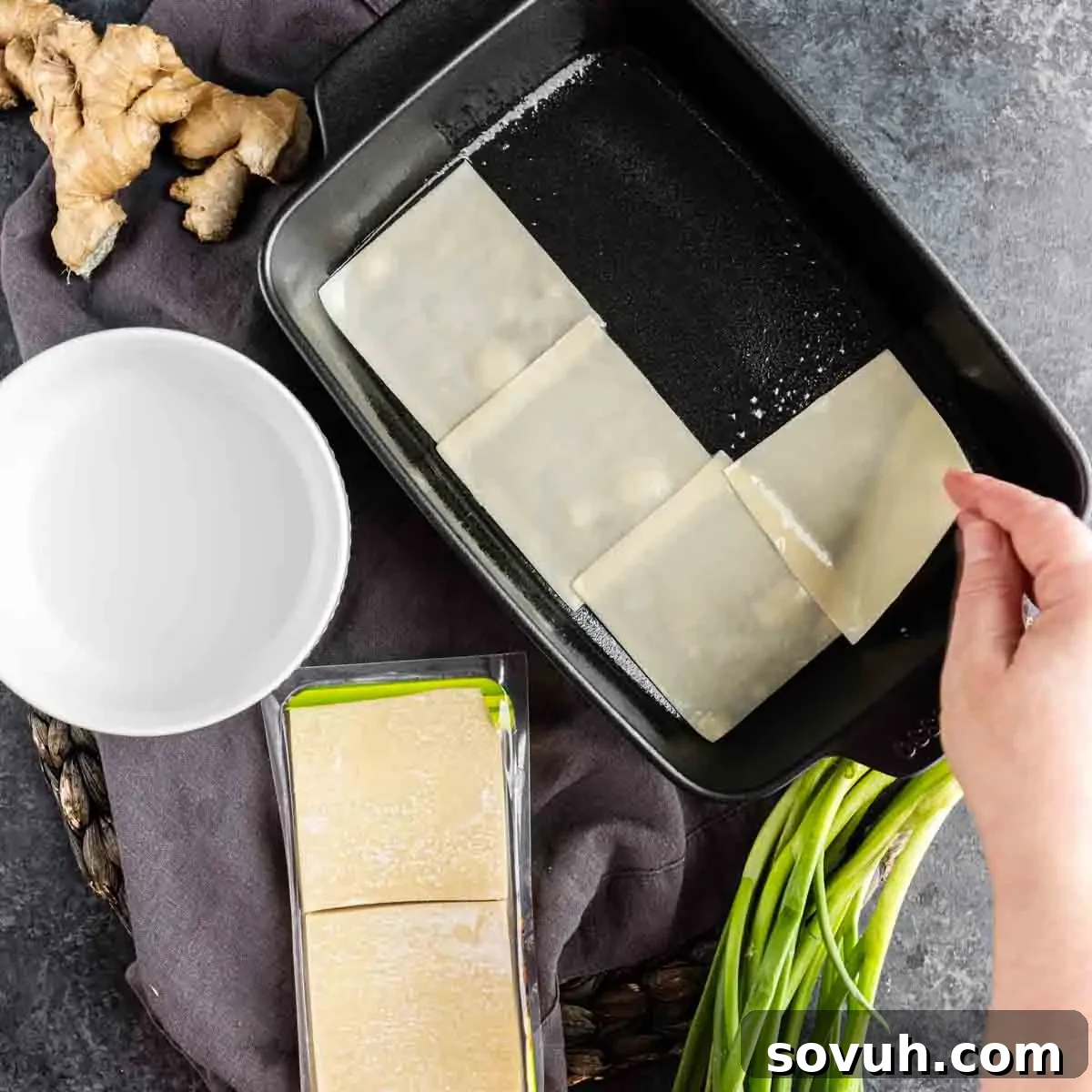 Viral Wonton Lasagna Twist 5 A hand places a wonton wrapper into a baking dish for a pork dumpling casserole; nearby are green onions, ginger, a bowl of water, and a pack of wonton wrappers—a nod to the viral dumpling lasagna trend.