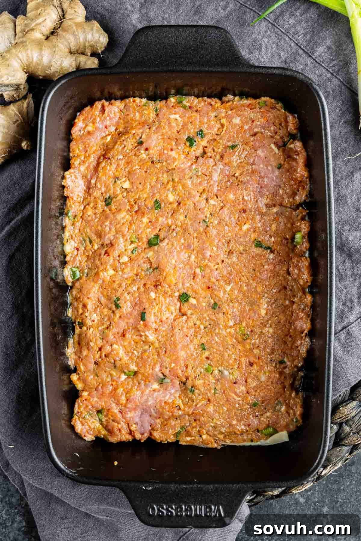Viral Wonton Lasagna Twist 6 A rectangular black baking dish filled with seasoned, uncooked ground meat mixture for a pork dumpling casserole, placed on a dark cloth next to ginger root and green onions.