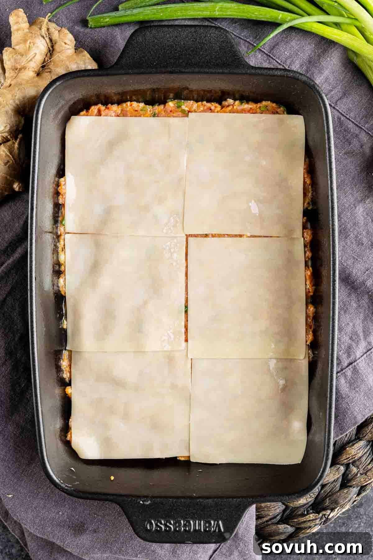 Viral Wonton Lasagna Twist 7 A black rectangular baking dish filled with layered noodles and filling, topped with six uncooked lasagna sheets, resembles a creative dumpling lasagna. Green onions and ginger sit beside the dish, perfect for a TikTok dumpling lasagna recipe.