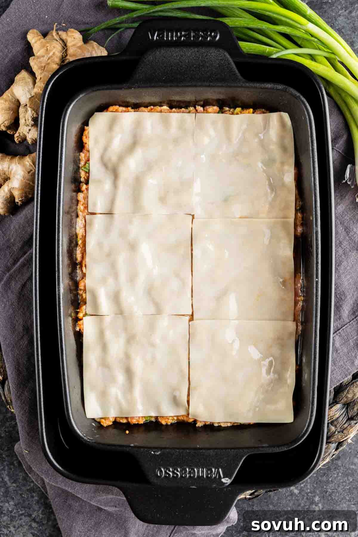 Viral Wonton Lasagna Twist 8 A rectangular baking dish filled with a layered casserole, topped with six sheets of uncooked pasta, hints at a viral dumpling lasagna inspired by TikTok. Green onions and ginger root are visible nearby.