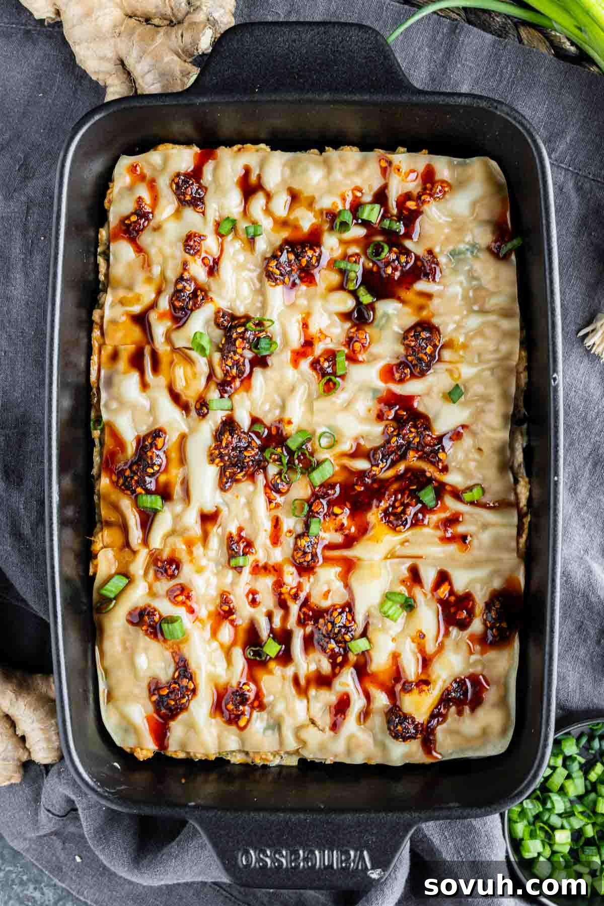 Viral Wonton Lasagna Twist 10 Rectangular baking dish filled with a pork dumpling casserole topped with sauces, chopped green onions, and chili flakes, placed on a dark cloth.