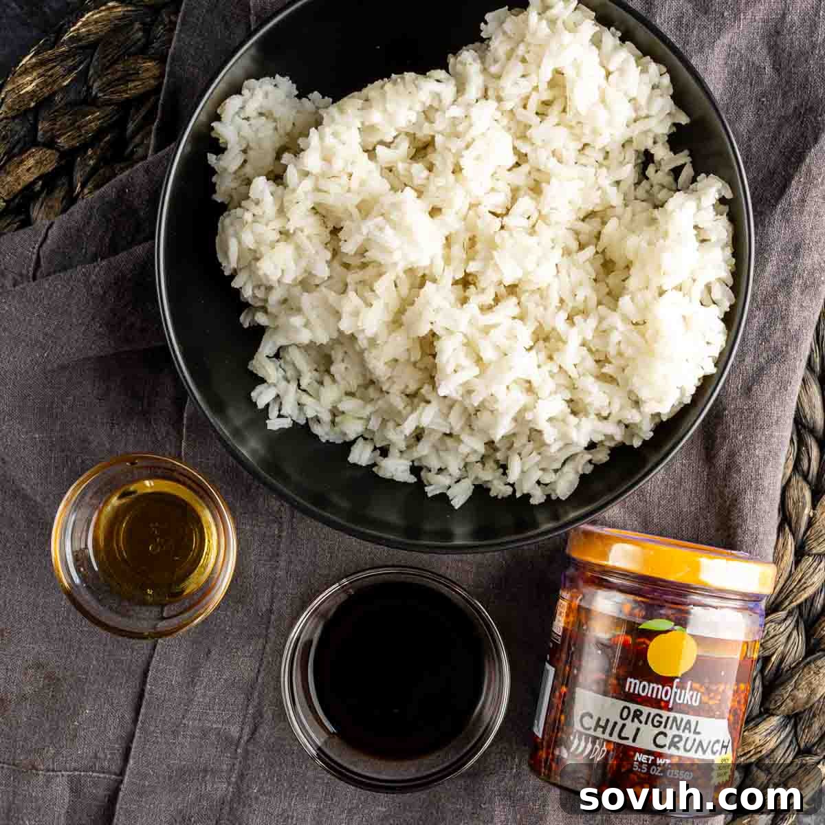 A bowl of cooked white rice next to a jar of Momofuku Chili Crunch, a small glass of sesame oil, and a small bowl of soy sauce—essential ingredients for the viral TikTok crispy rice recipe.