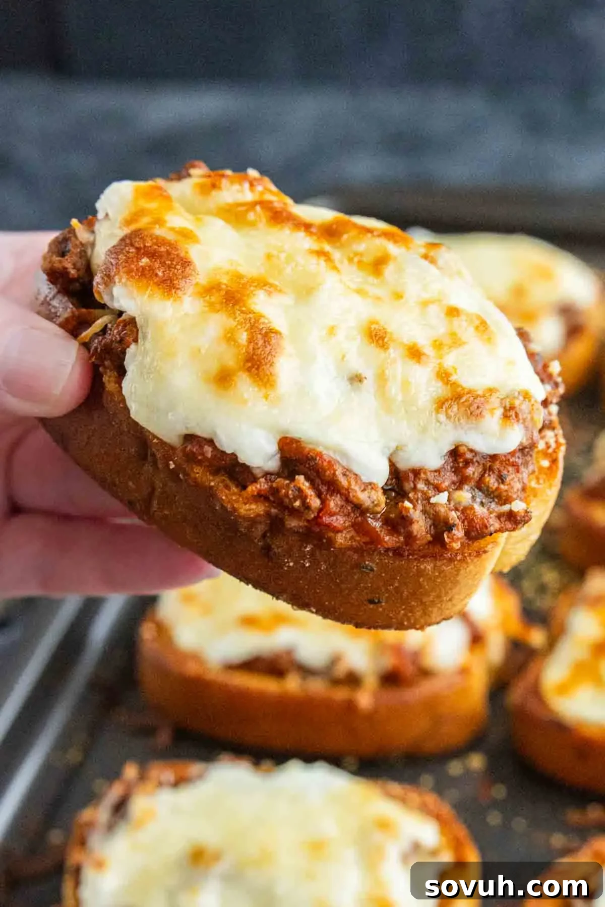 Texas Toast Garlic Bread Lasagna Recipe: Cheesy Weeknight Dinner 6 A hand holds a slice of easy garlic bread lasagna, topped with ground meat and melted cheese; more slices are visible on a tray in the background.