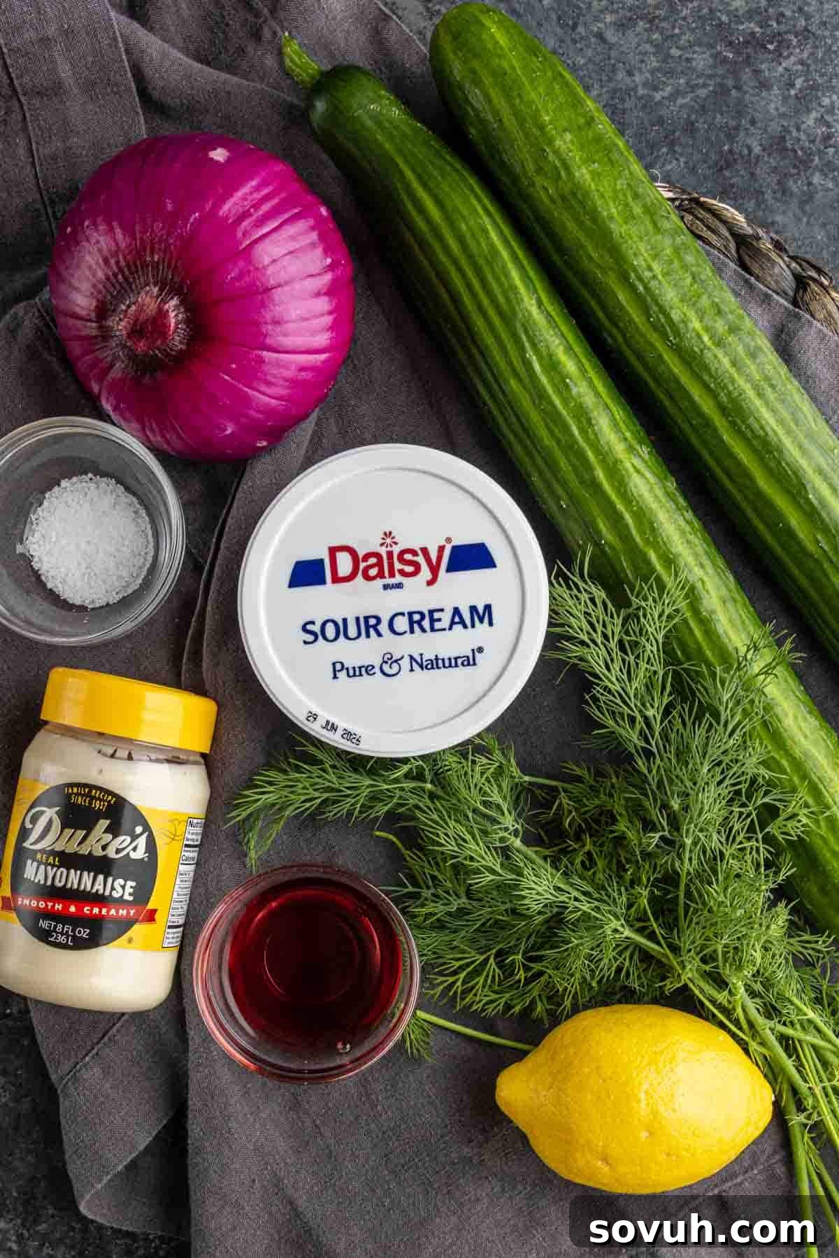 Creamy Summer Cucumber Salad with Dill and Yogurt 3 Top-down view of ingredients for creamy cucumber salad on a cloth: red onion, cucumbers, fresh dill, lemon, sour cream, mayonnaise, salt, and a small bowl of red liquid.