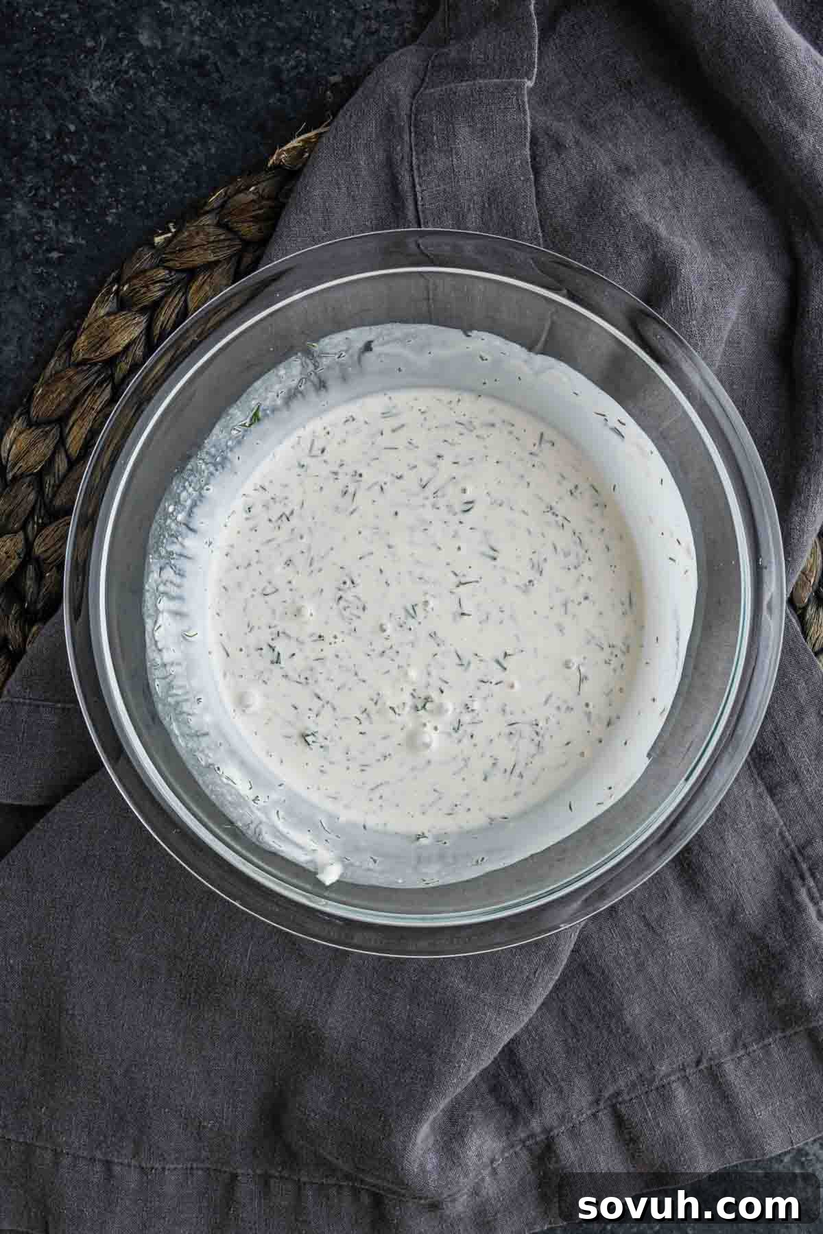 Creamy Summer Cucumber Salad with Dill and Yogurt 5 A glass bowl filled with creamy ranch dressing, perfect for a dill cucumber salad, sits on a dark gray cloth and woven mat.