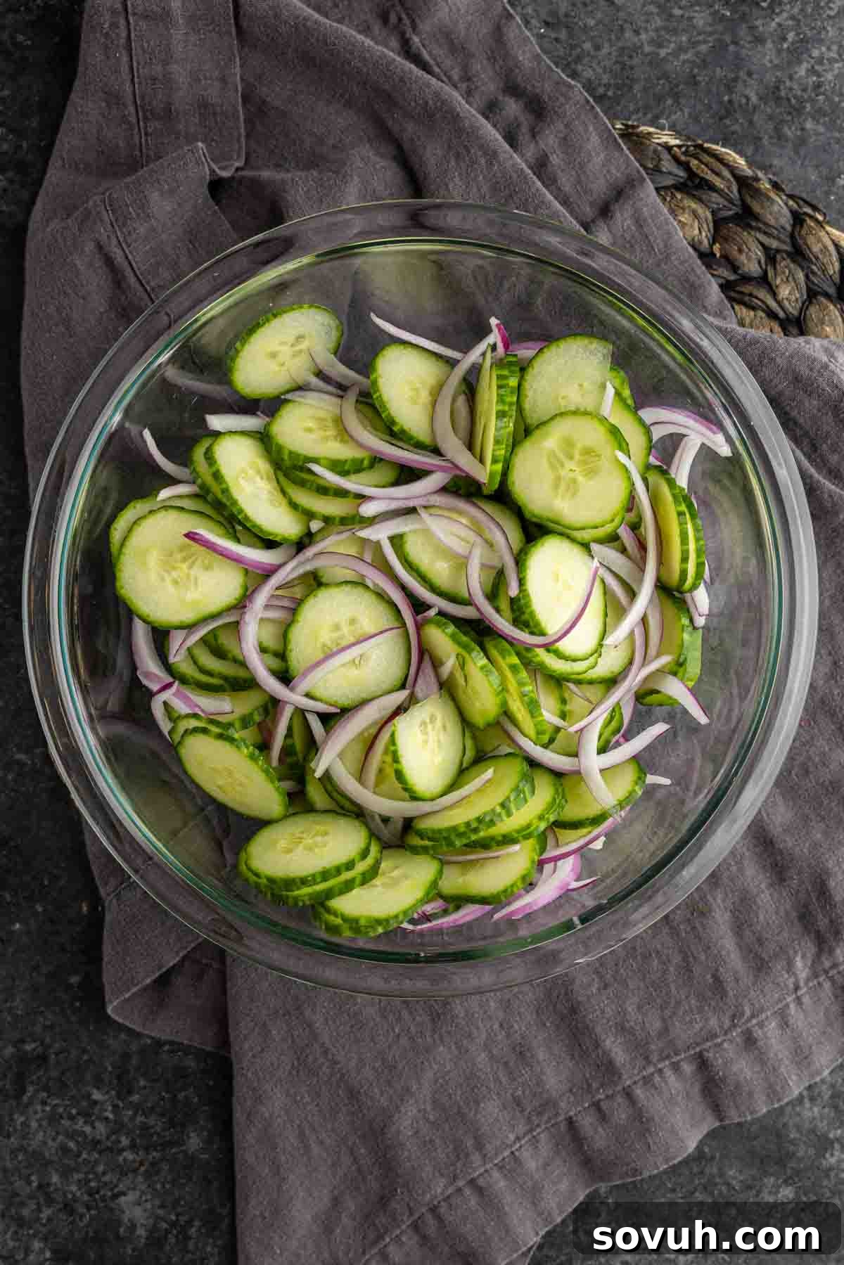 Creamy Summer Cucumber Salad with Dill and Yogurt 6 A glass bowl filled with sliced cucumbers and thinly sliced red onions, ready for a refreshing cucumber onion salad, sits on a dark cloth and countertop.