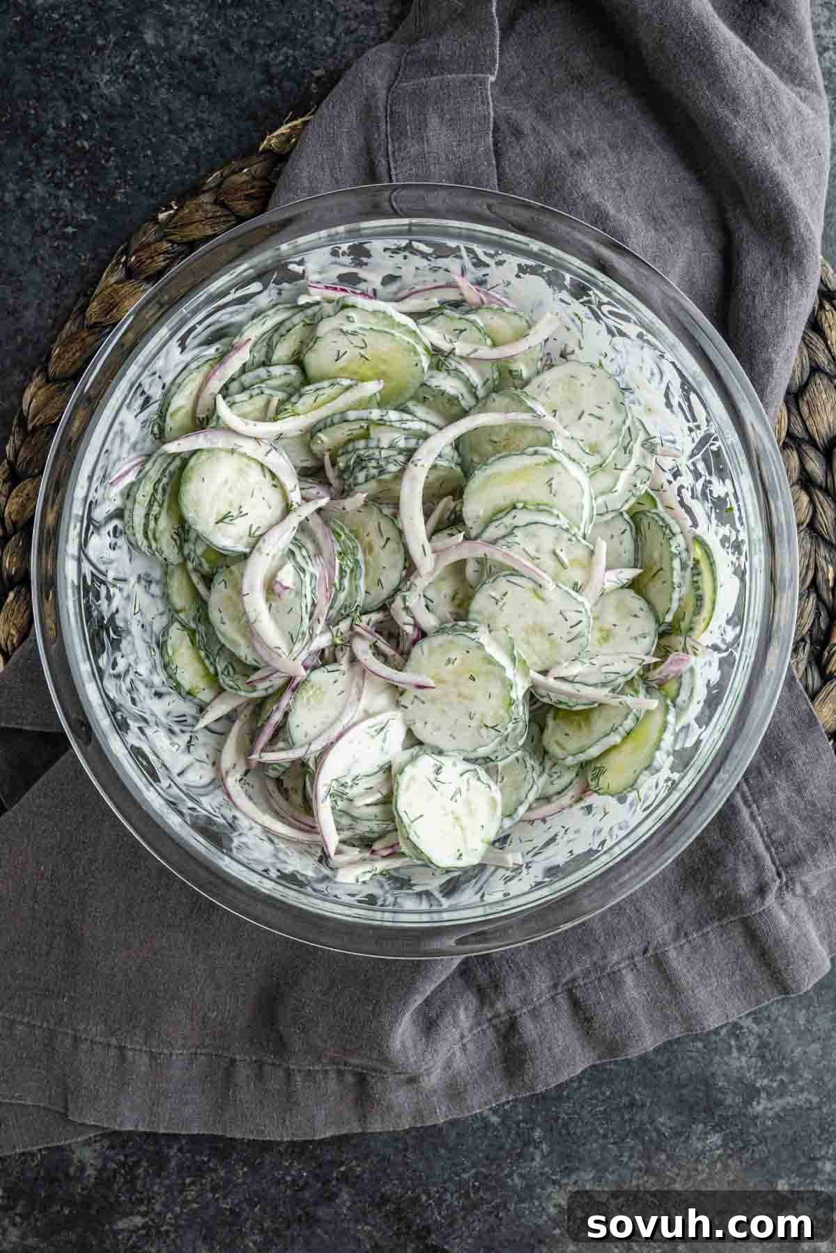 Creamy Summer Cucumber Salad with Dill and Yogurt 7 A glass bowl filled with creamy cucumber salad and red onions, garnished with dill, sits on a gray cloth and woven mat.