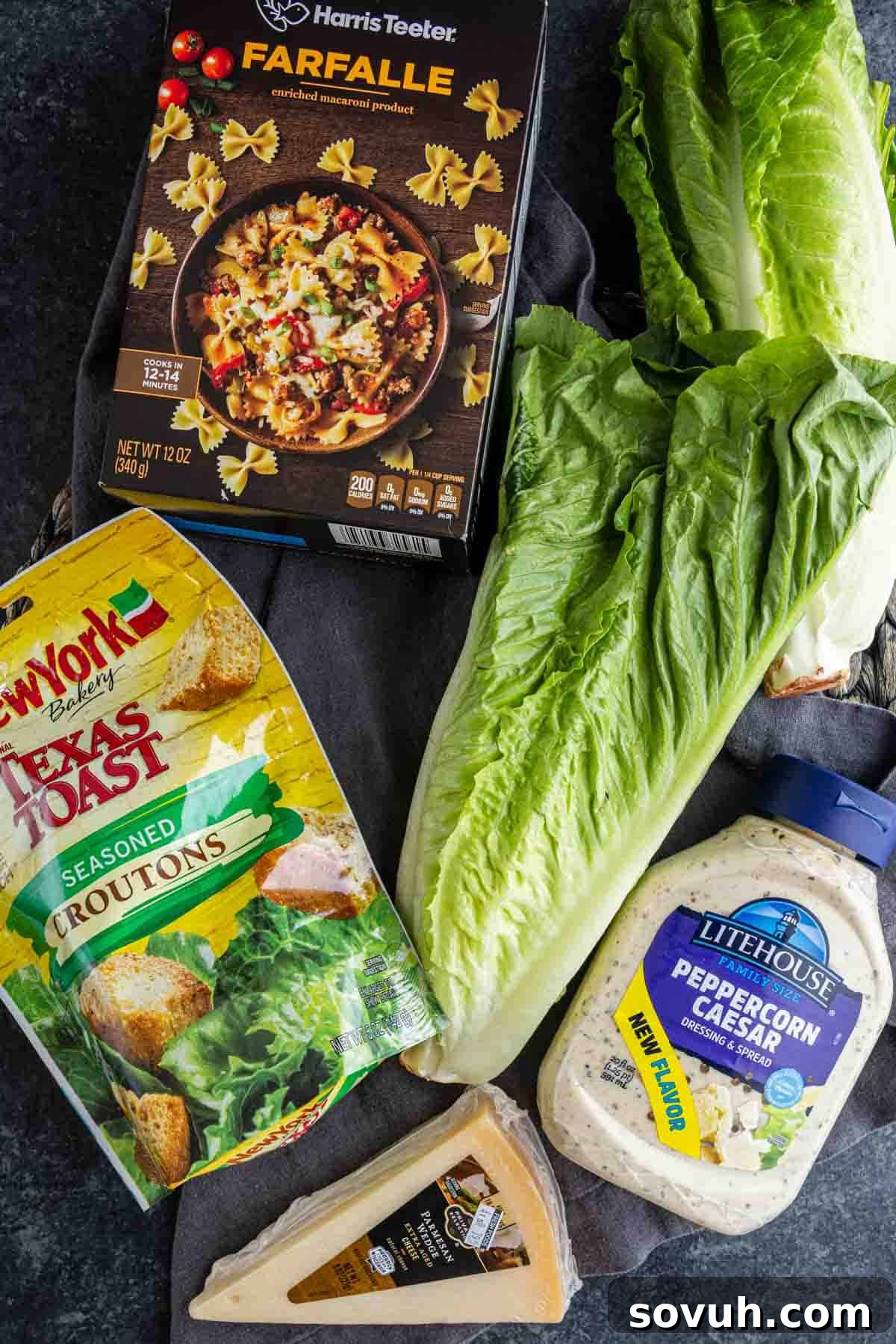 Creamy Caesar Pasta Salad Recipe for Summer Potlucks 3 A box of farfalle pasta, romaine lettuce, seasoned croutons, peppercorn Caesar dressing, and Parmesan—everything you need for a delicious Caesar pasta salad recipe on a dark surface.