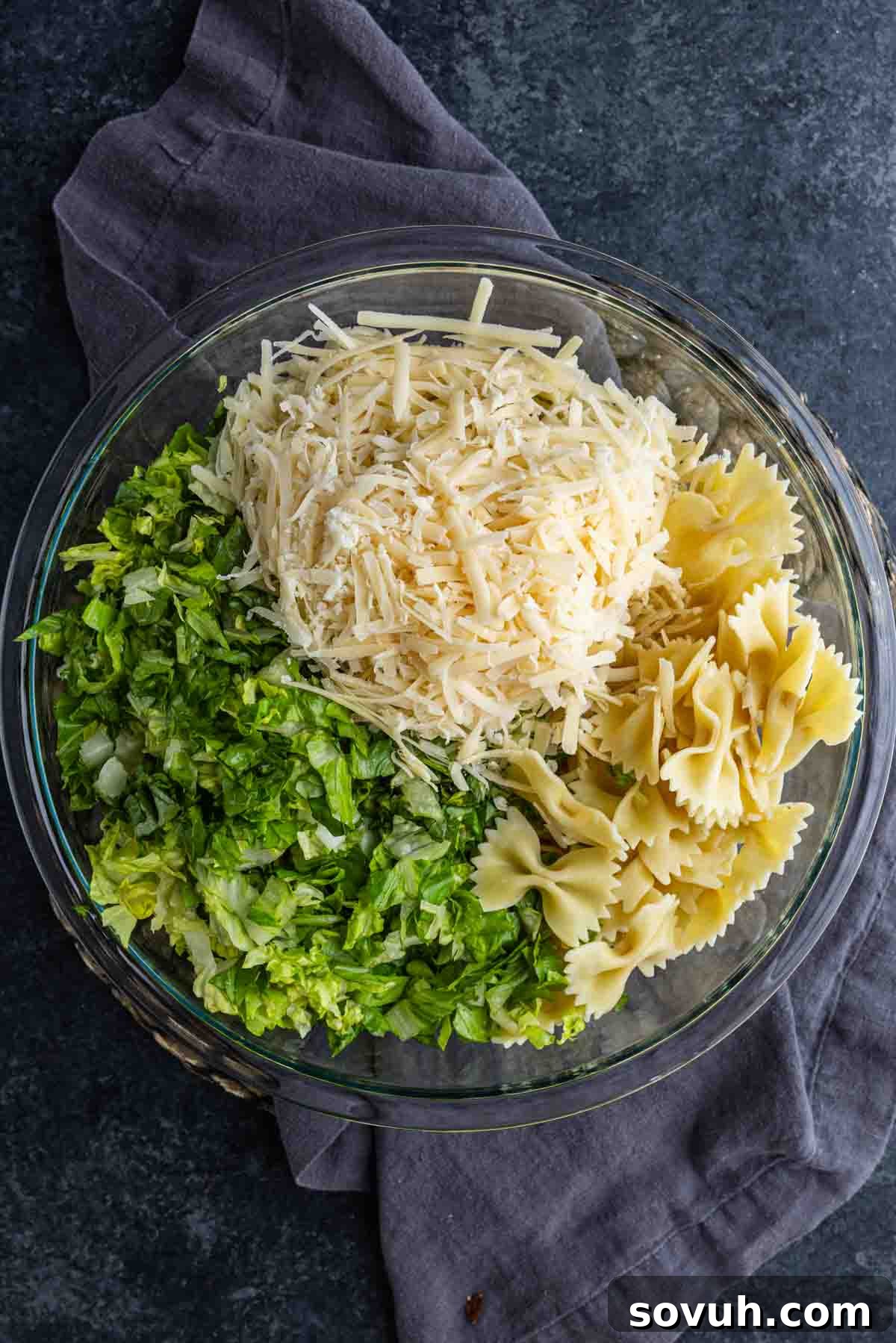 Creamy Caesar Pasta Salad Recipe for Summer Potlucks 4 A glass bowl containing a cold pasta salad with cooked bowtie pasta, shredded lettuce, and grated cheese, placed on a dark surface with a gray cloth underneath.