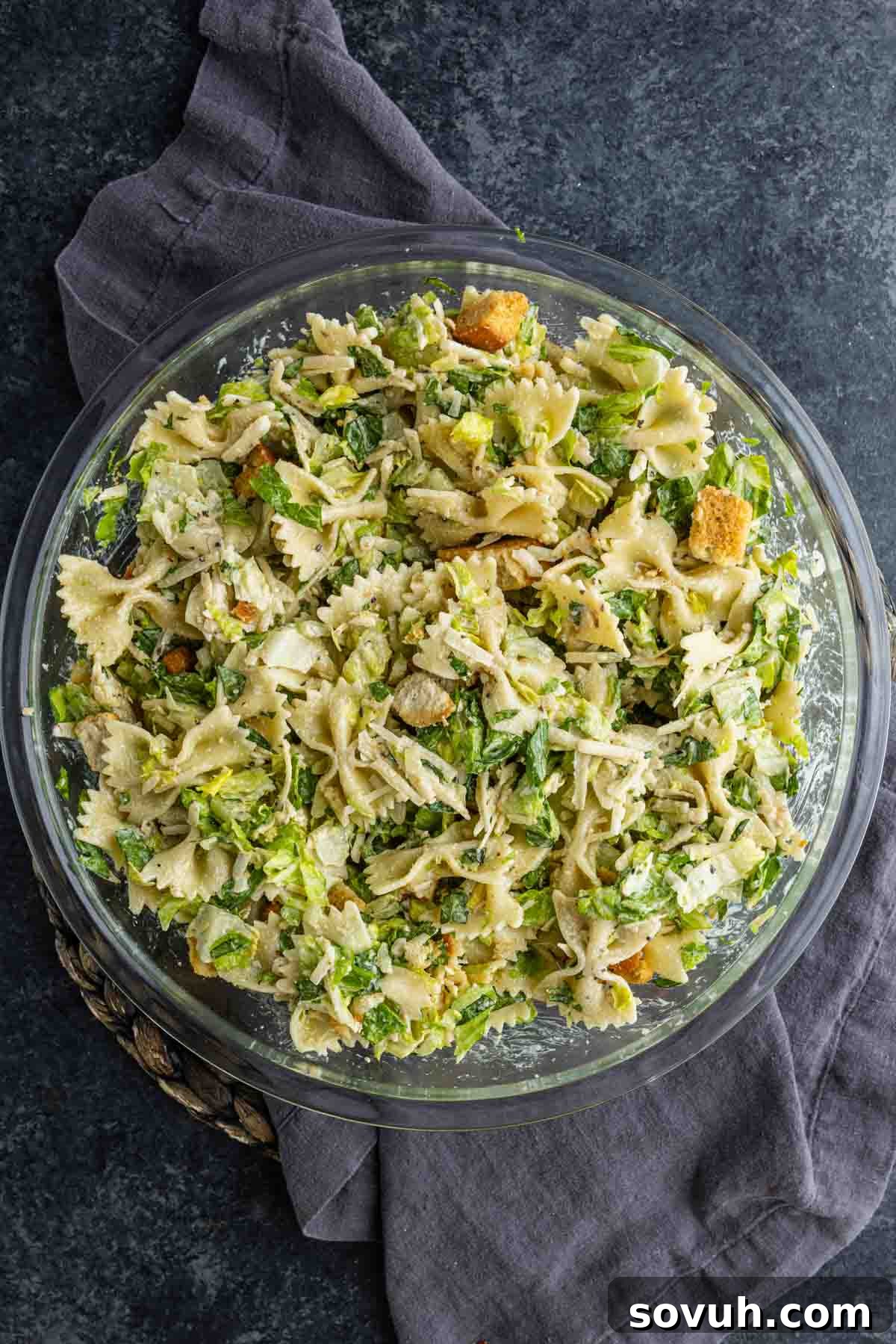 Creamy Caesar Pasta Salad Recipe for Summer Potlucks 5 A glass bowl filled with an easy pasta salad of bowtie pasta, chopped lettuce, shredded chicken, croutons, and cheese sits on a dark surface with a gray cloth.