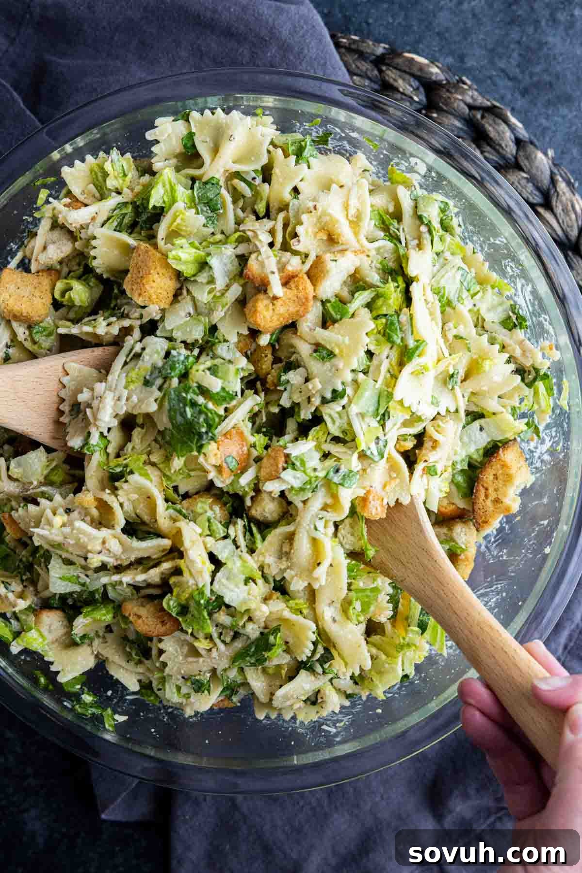 Creamy Caesar Pasta Salad Recipe for Summer Potlucks 6 A glass bowl filled with Caesar pasta salad—bowtie pasta mixed with lettuce, croutons, shredded cheese, and creamy dressing—being tossed with wooden utensils for the perfect cold pasta salad.