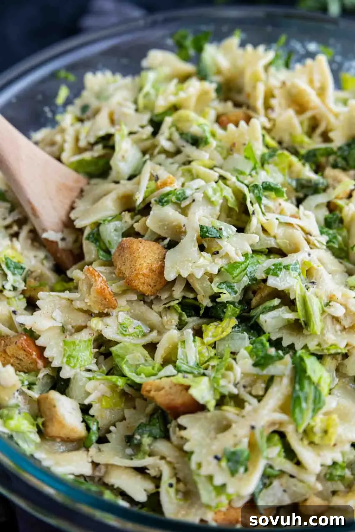 Creamy Caesar Pasta Salad Recipe for Summer Potlucks 7 A wooden spoon in a bowl of Caesar salad pasta salad with bowtie pasta, lettuce, croutons, and a creamy dressing—an easy pasta salad perfect for any occasion.