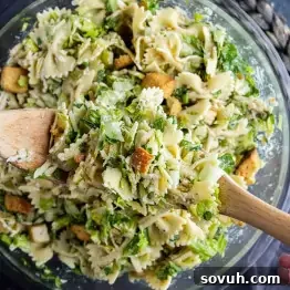 Creamy Caesar Pasta Salad Recipe for Summer Potlucks 8 A bowl of Caesar salad pasta salad with farfalle, chopped greens, croutons, and grated cheese is being mixed with wooden utensils for an easy pasta salad perfect for any meal.