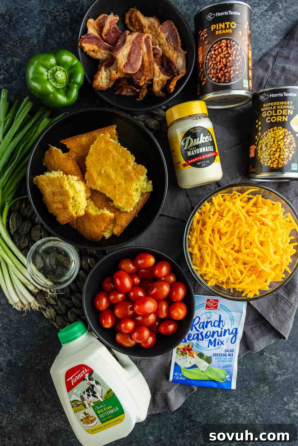 Southern Cornbread Salad Recipe for Potlucks and BBQs 3 Ingredients for a cornbread salad with ranch dressing arranged on a dark surface, perfect as a summer BBQ side—cornbread, bacon, cheddar, grape tomatoes, ranch mix, mayo, pinto beans, corn, green onions, green pepper, and buttermilk.