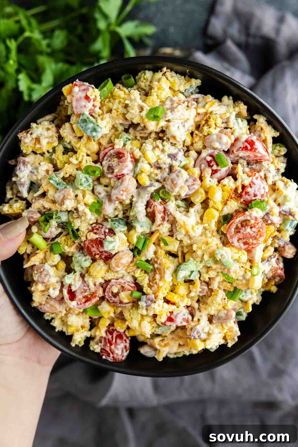 Southern Cornbread Salad Recipe for Potlucks and BBQs 5 A bowl of salad with cherry tomatoes, corn, chopped green onions, and creamy dressing is held by a hand—perfect as a Southern potluck side dish. Fresh parsley adds color in the background, making it ideal for any summer BBQ side.
