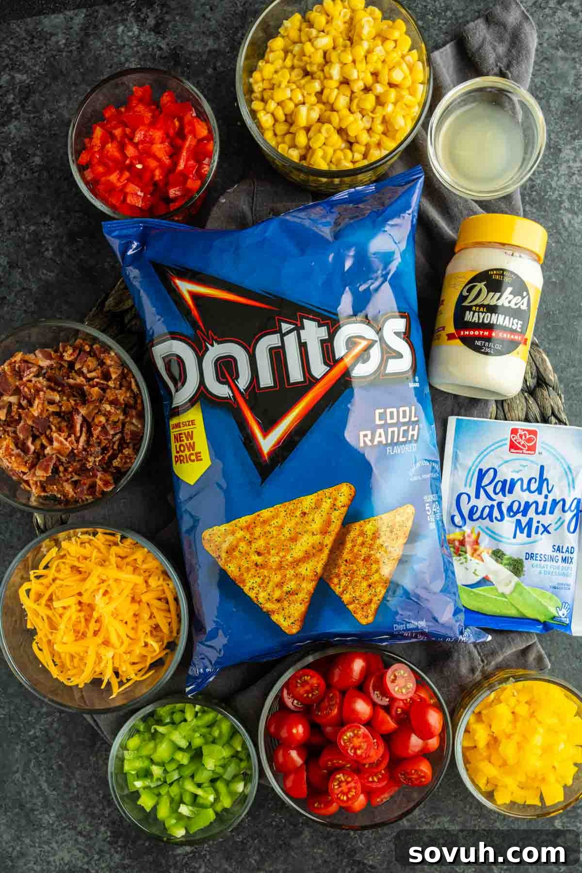 Crunchy Cool Ranch Doritos Salad with Bacon & Sweet Corn 3 A bag of Cool Ranch Doritos is surrounded by bowls of chopped red pepper, corn, bacon, cheddar cheese, celery, tomatoes, green pepper, mayonnaise, and a packet of ranch seasoning—everything you need for a flavorful Cool Ranch Doritos Salad.