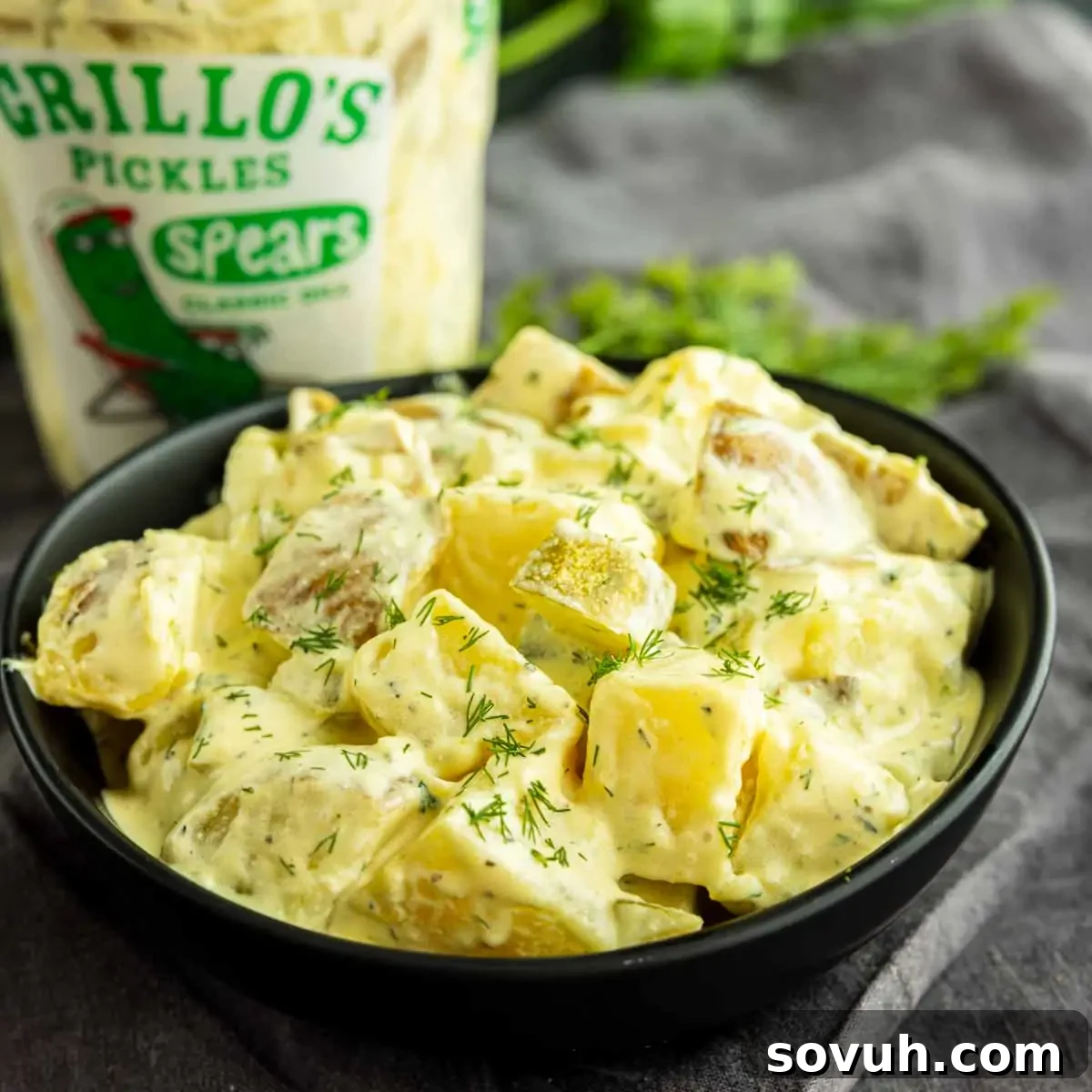 Creamy Dill Pickle Potato Salad with Tangy Dressing 2 A bowl of creamy dill pickle potato salad sits in front of a container of Grillo's Pickles spears, garnished with fresh dill.