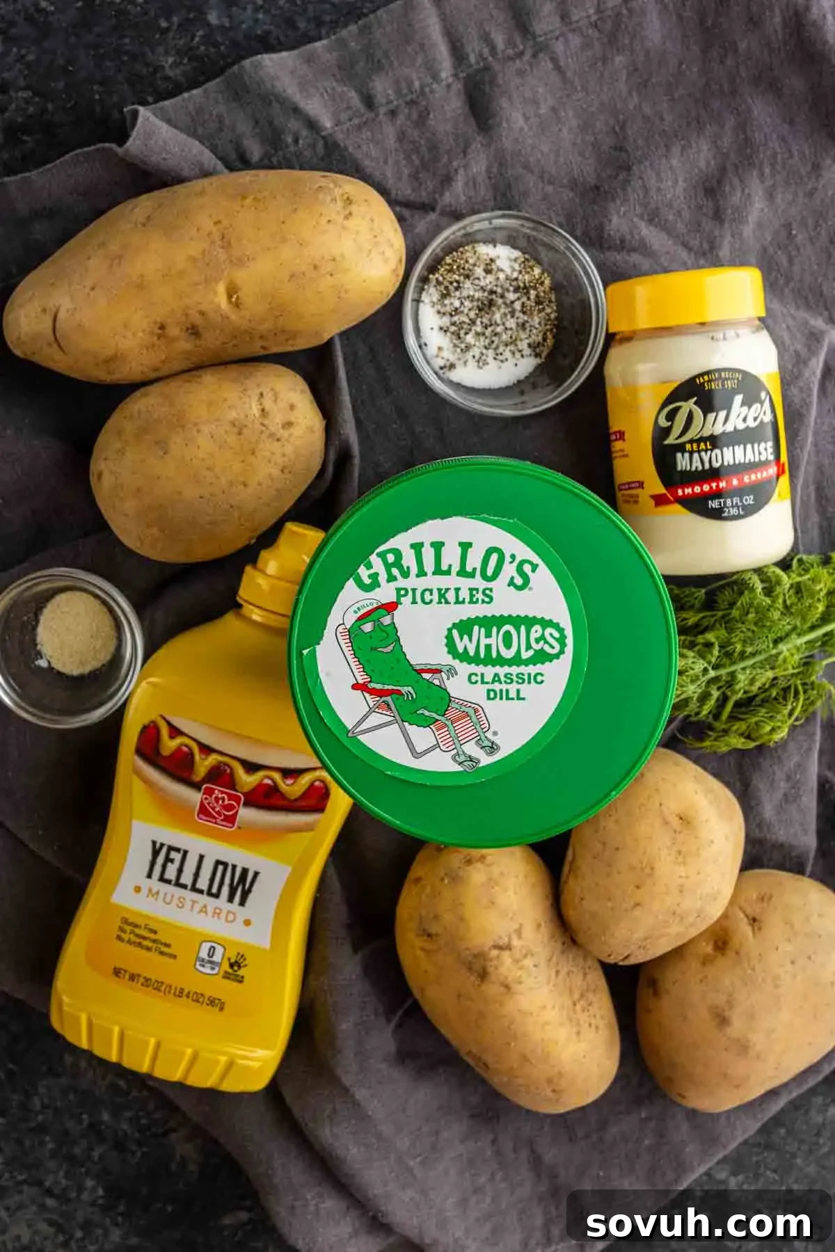 Creamy Dill Pickle Potato Salad with Tangy Dressing 3 Four russet potatoes, a jar of mayonnaise, yellow mustard, dill pickles, fresh dill, ground pepper, and spices are arranged on a dark cloth—perfect for preparing a flavorful make-ahead dill pickle potato salad.
