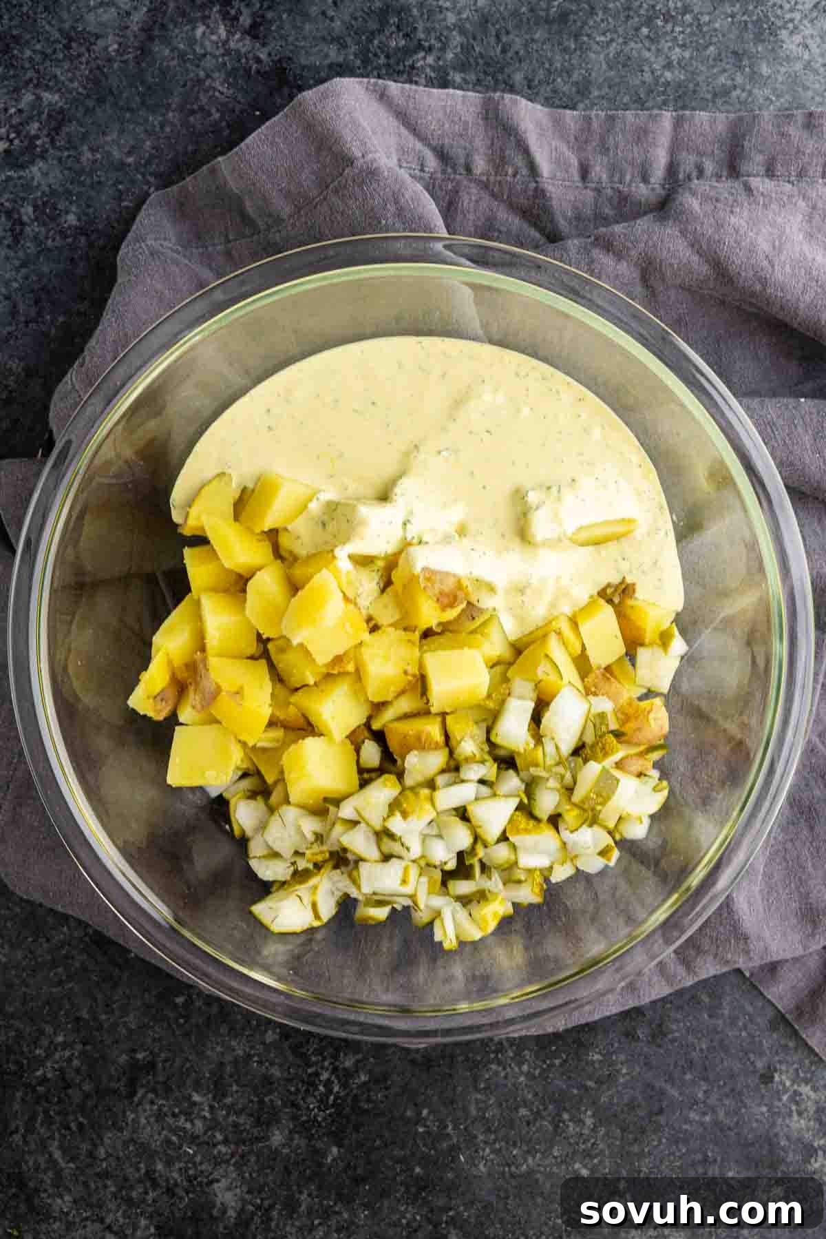 Creamy Dill Pickle Potato Salad with Tangy Dressing 5 A glass bowl holds a creamy yellow pickle potato salad with chopped potatoes, diced onion, and pickles, all set atop a gray cloth.