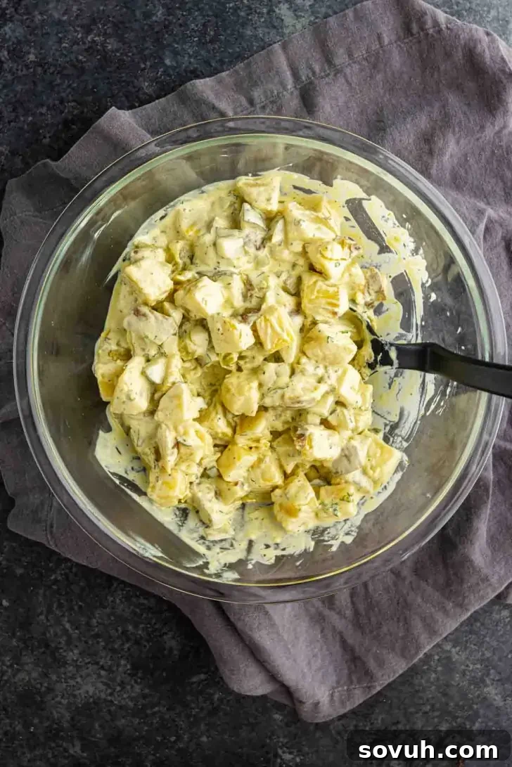 Creamy Dill Pickle Potato Salad with Tangy Dressing 6 A glass bowl filled with creamy dill pickle potato salad sits on a folded gray cloth, with a black serving spoon inside the bowl.
