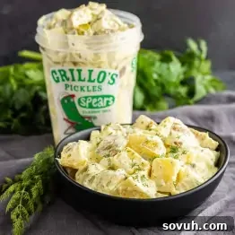 Creamy Dill Pickle Potato Salad with Tangy Dressing 9 A bowl of creamy dill pickle potato salad, garnished with dill, sits in front of a container of Grillo’s Pickles spears, with fresh herbs in the background.