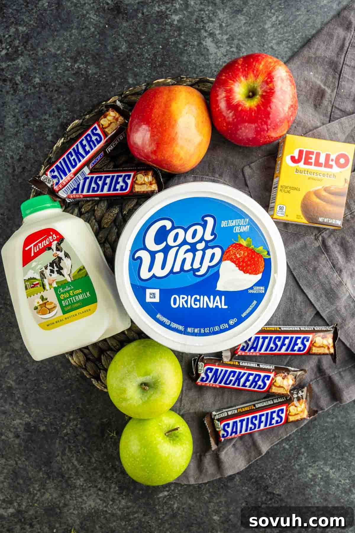 Snickers Apple Dessert Salad Recipe | Chocolate Caramel Apple Fluff 3 A classic Snickers Salad recipe: four Snickers bars, two red apples, two green apples, a carton of buttermilk, Cool Whip, and a box of Jello butterscotch pudding mix on a dark surface—everything you need for an easy Snickers Salad.
