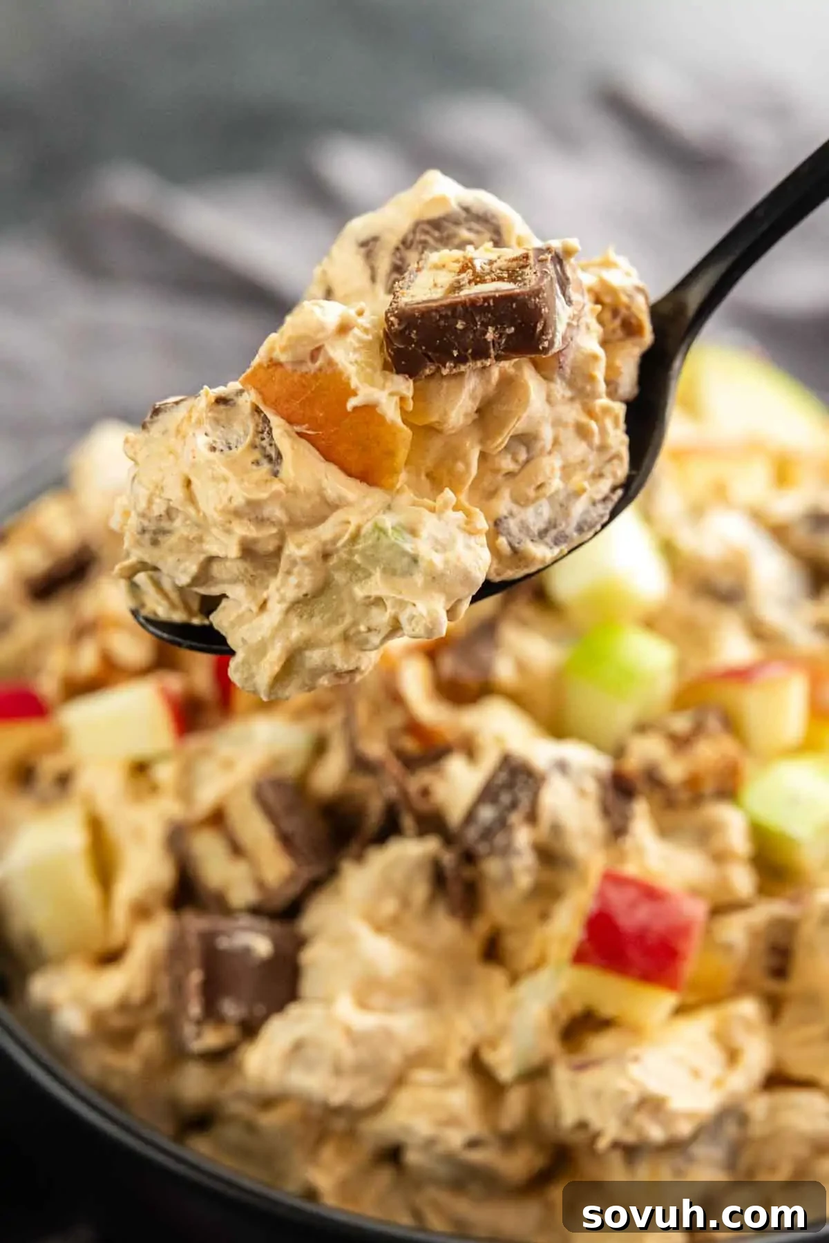 Snickers Apple Dessert Salad Recipe | Chocolate Caramel Apple Fluff 5 A close-up of a spoonful of creamy Snickers Apple Salad with chopped apples and chocolate pieces, held above a bowl filled with the same mixture.