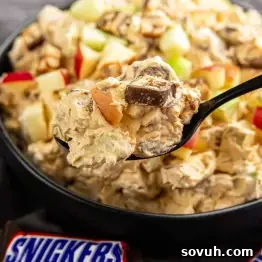 Snickers Apple Dessert Salad Recipe | Chocolate Caramel Apple Fluff 7 A spoonful of creamy Snicker Apple Salad with chopped apples and pieces of Snickers bar held above a bowl of the same salad; a Snickers wrapper is visible in the foreground.