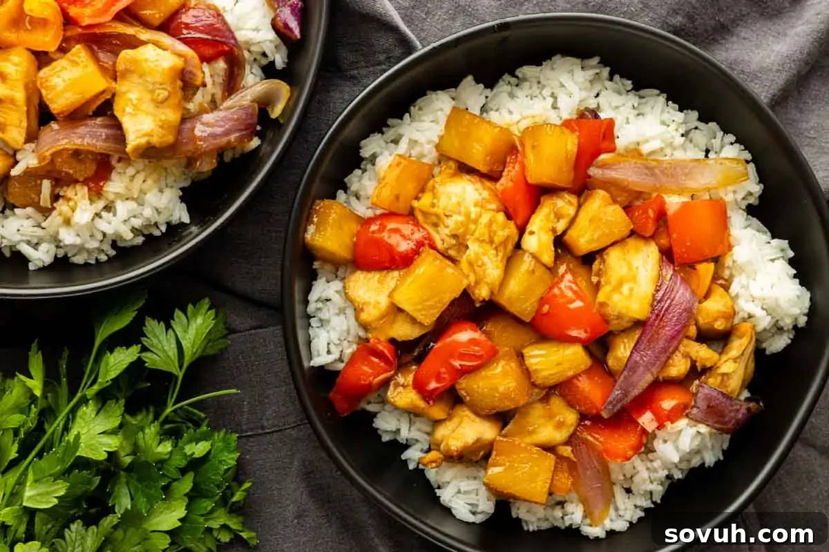 Sheet Pan Hawaiian Chicken with Pineapple and Teriyaki Glaze 2 A bowl of white rice topped with sweet and sour chicken, bell peppers, pineapple chunks, and red onion—this one pan Hawaiian chicken makes for an easy meal. Another partial bowl and fresh parsley are next to it.