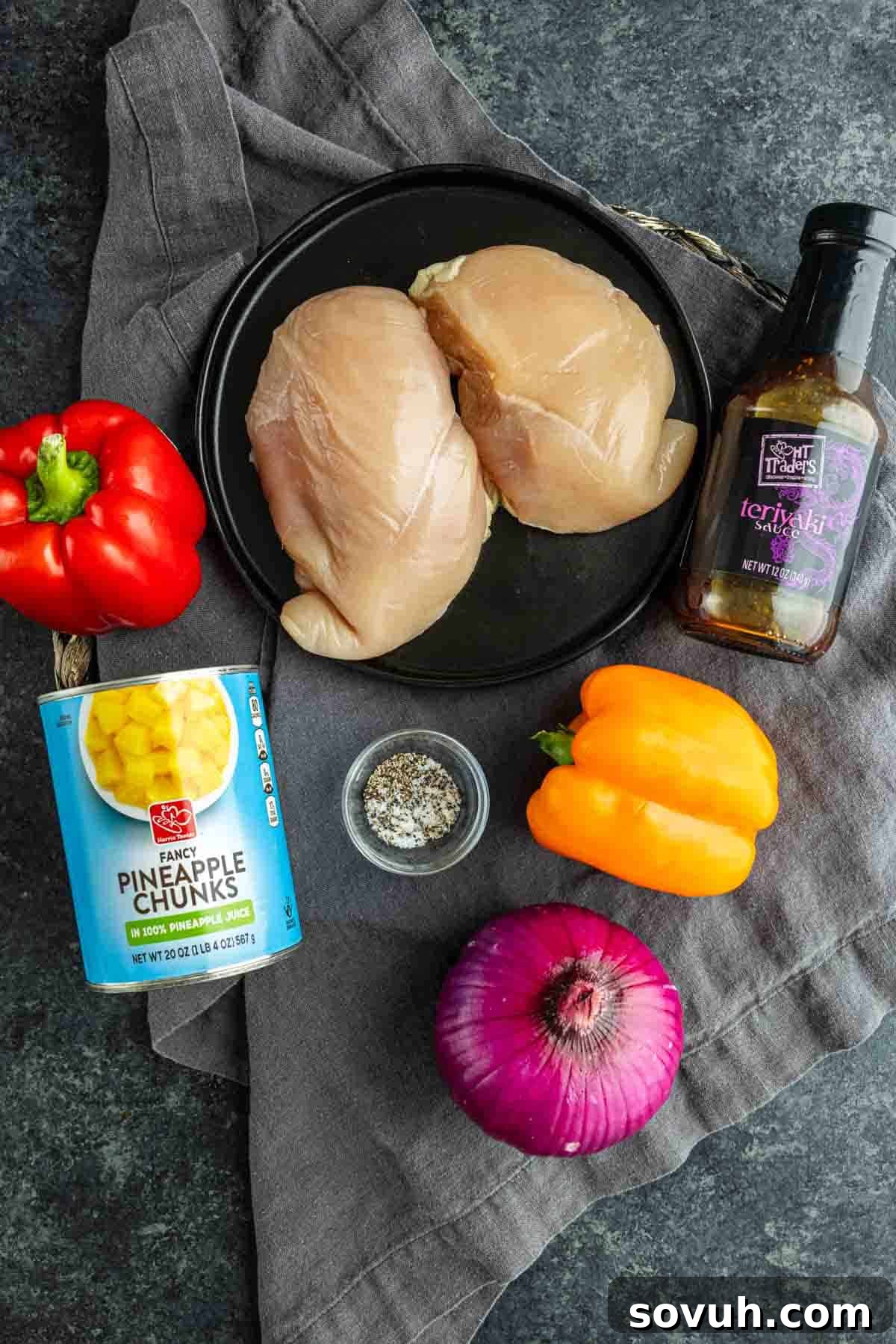 Sheet Pan Hawaiian Chicken with Pineapple and Teriyaki Glaze 3 Raw chicken breasts on a black plate, surrounded by red and yellow bell peppers, red onion, pineapple chunks, seasoning blend, and teriyaki sauce—perfect for an easy weeknight dinner like sheet pan Hawaiian chicken.