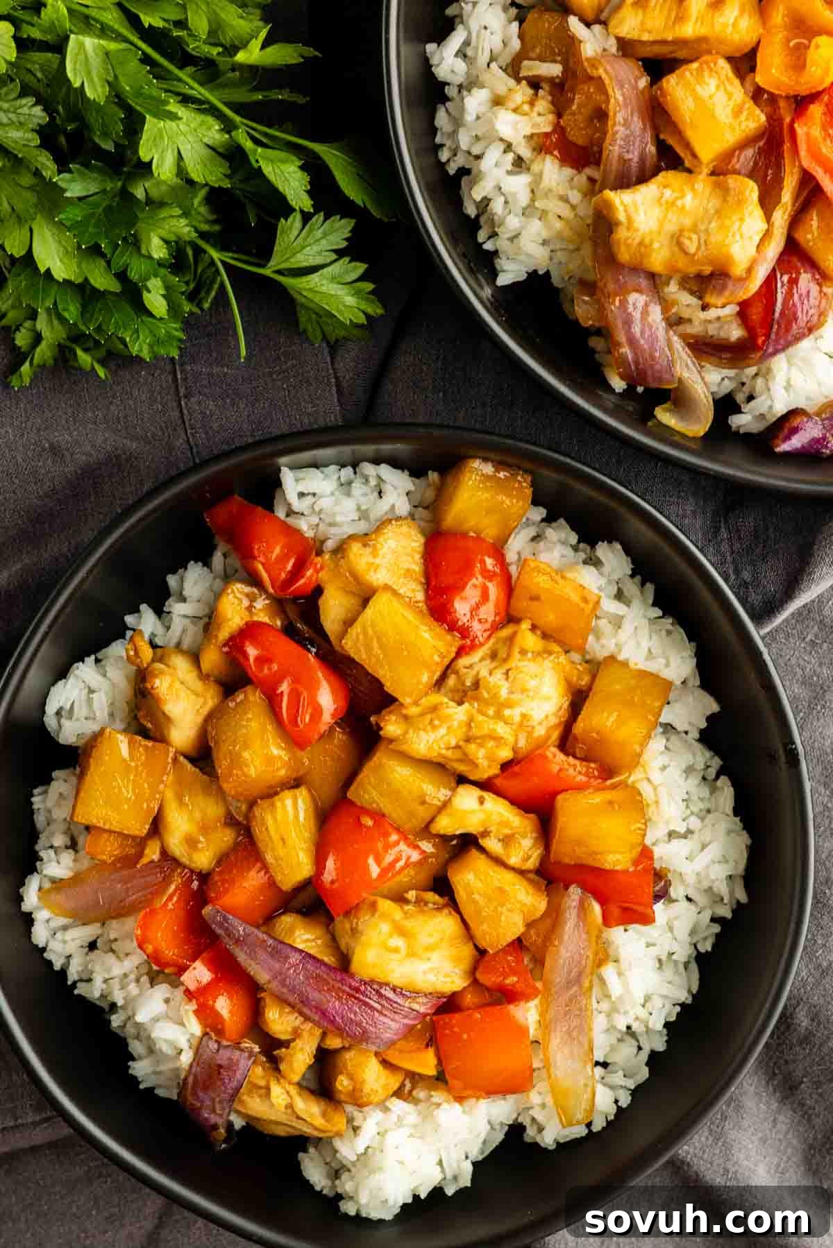 Sheet Pan Hawaiian Chicken with Pineapple and Teriyaki Glaze 5 Two bowls of white rice topped with one pan Hawaiian chicken—stir-fried with red bell peppers, pineapple chunks, and red onions—sit on a dark tablecloth beside fresh parsley. An easy weeknight dinner that's bursting with tropical flavor.