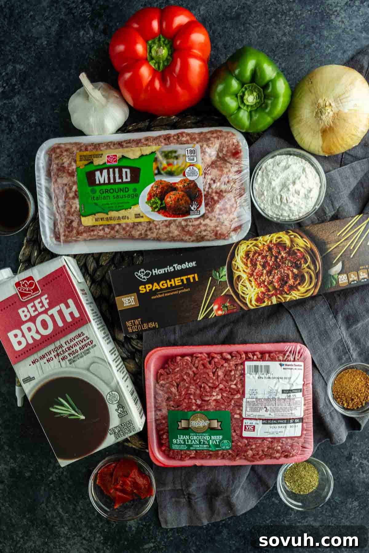 Viral Cajun Dirty Spaghetti Recipe for Dinner That Delights 3 Top-down view of ingredients for Cajun spaghetti, including ground beef, Italian sausage, spaghetti pasta, bell peppers, onion, garlic, beef broth, tomato paste, and spices on a dark surface.