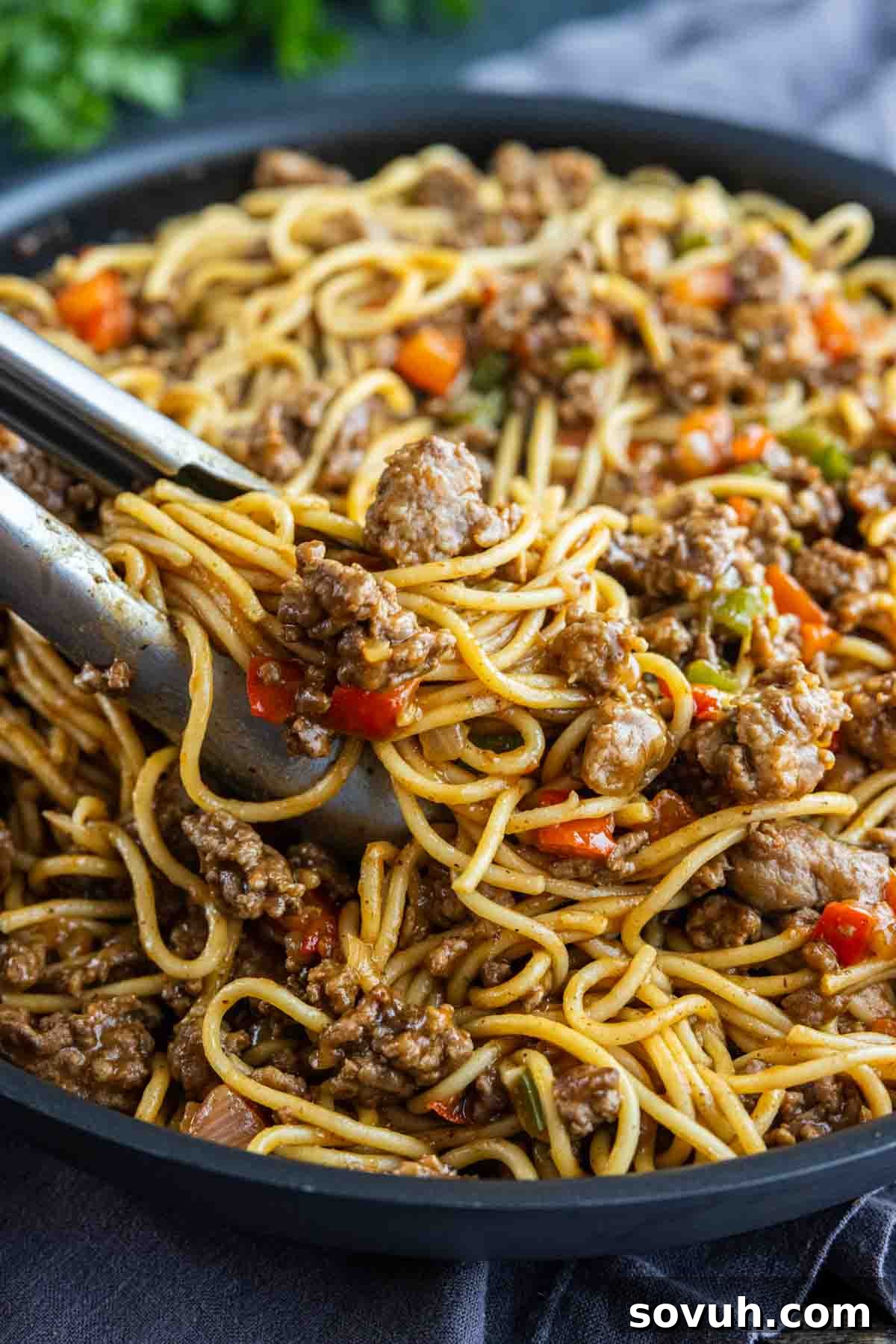 Viral Cajun Dirty Spaghetti Recipe for Dinner That Delights 5 Close-up of tongs lifting stir-fried noodles with ground beef and mixed vegetables from a skillet, inspired by viral dirty spaghetti for a flavorful twist.