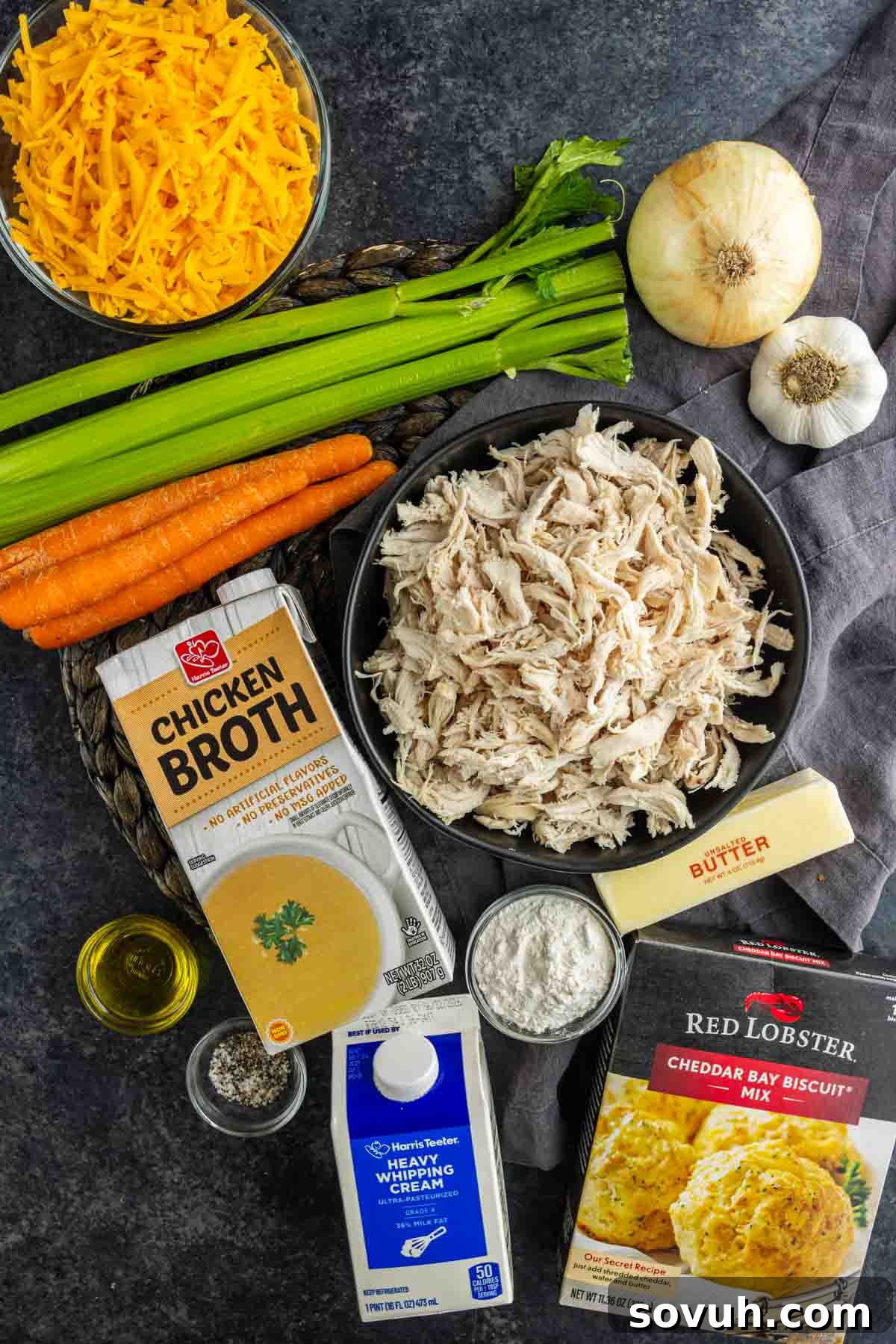 Ingredients for a chicken cobbler arranged on a dark surface, featuring shredded chicken, cheddar cheese, celery, carrots, onion, garlic, chicken broth, cream, butter, oil, flour and biscuit mix. Perfect for a Cheddar Bay Biscuit Chicken Casserole.