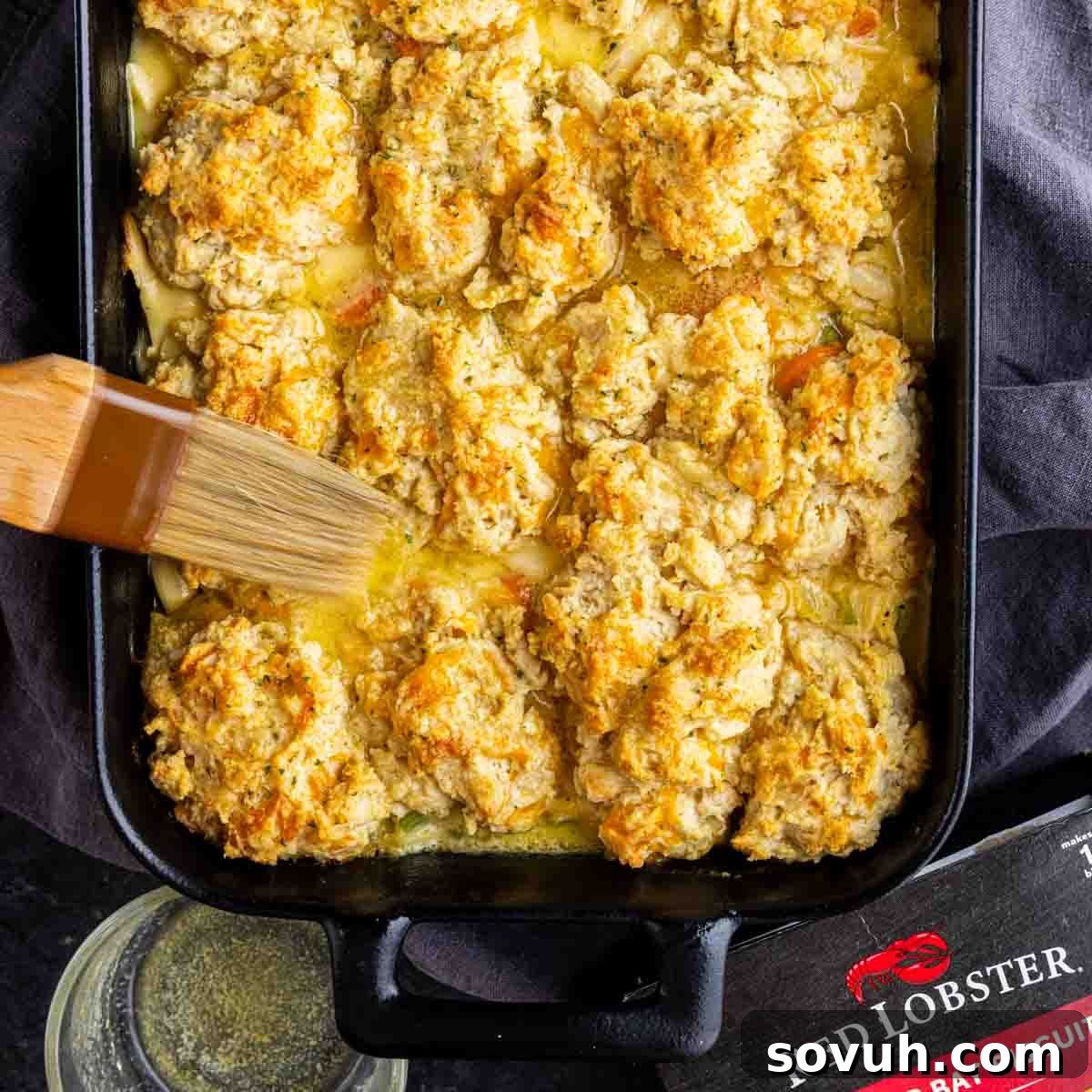 A baking dish filled with golden-brown Cheddar Bay Biscuit Chicken Casserole is being brushed with melted butter; a Red Lobster biscuit mix box is partially visible.