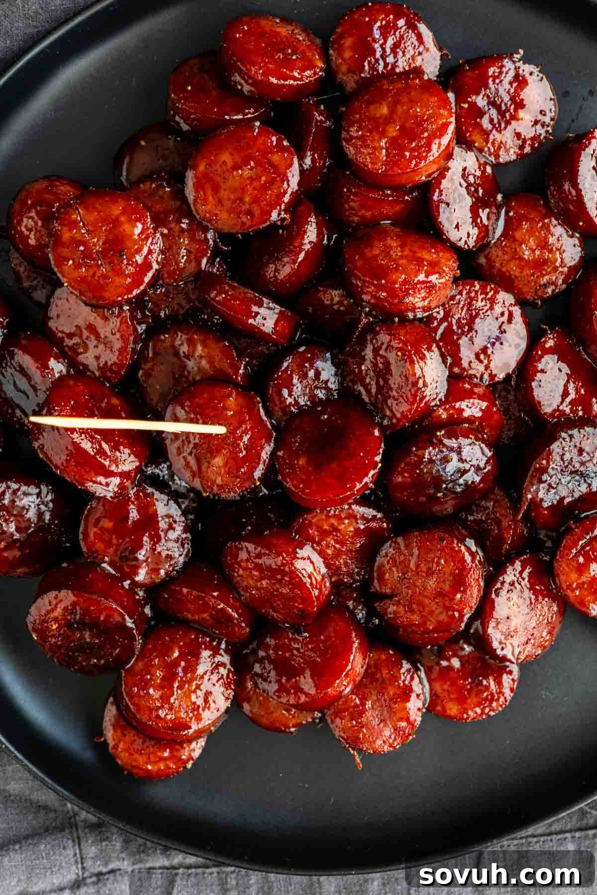 Oven-Baked Brown Sugar Barbecue Kielbasa Recipe 3 Sliced sausage pieces coated in a glossy Barbecue Brown Sugar Kielbasa sauce are arranged on a black plate, with a toothpick placed among them.