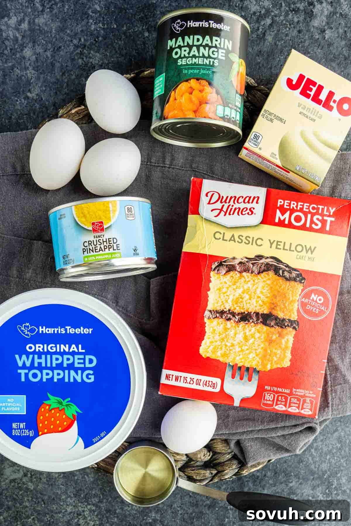 Southern Pig Pickin’ Cake with Mandarin Orange Glaze Recipe 3 Ingredients for a Pig Pickin’ Cake, including eggs, whipped topping, crushed pineapple, mandarin oranges, vanilla pudding mix, and classic yellow cake mix, arranged on a dark surface.