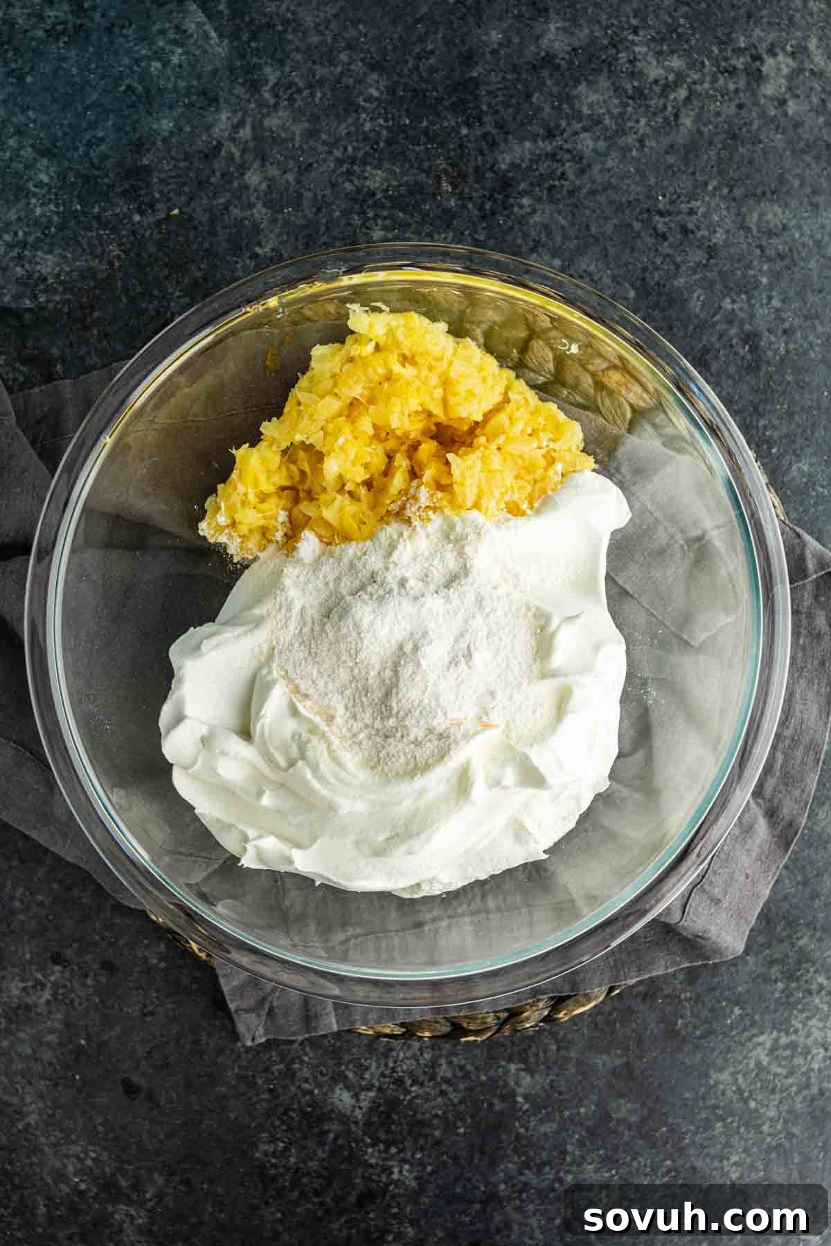 Southern Pig Pickin’ Cake with Mandarin Orange Glaze Recipe 4 A glass bowl containing crushed pineapple, whipped topping, and powdered pudding mix—a classic combination for a Southern dessert like the famous Pig Pickin’ Cake—sits on a gray napkin over a dark surface.