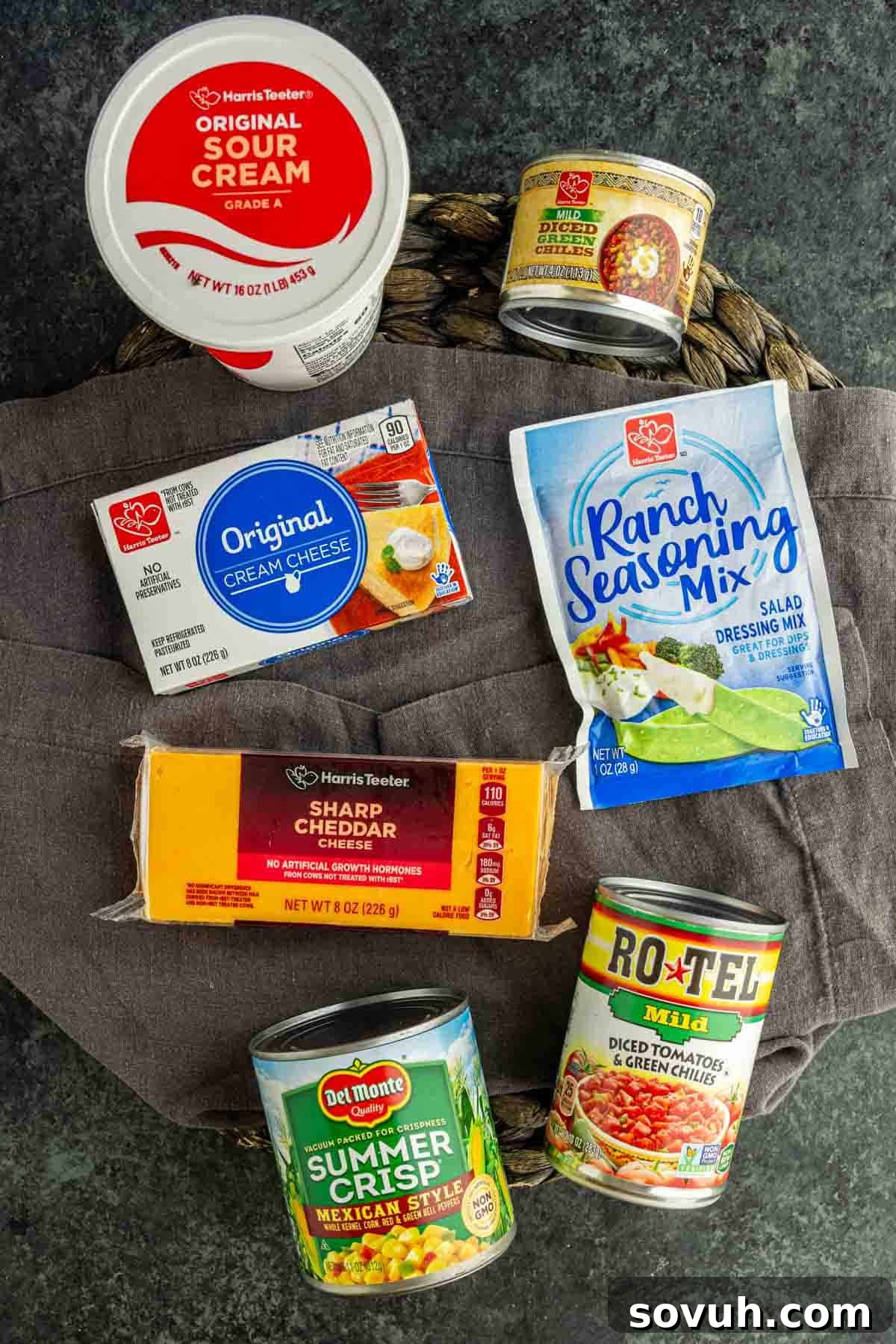 How to Make the Viral Boat Dip Everyone Loves 3 A variety of packaged food items, perfect for making cold corn dip or the viral boat dip, including sour cream, cream cheese, sharp cheddar cheese, ranch seasoning mix, summer crisp corn, and diced tomatoes with green chilies on a gray surface.