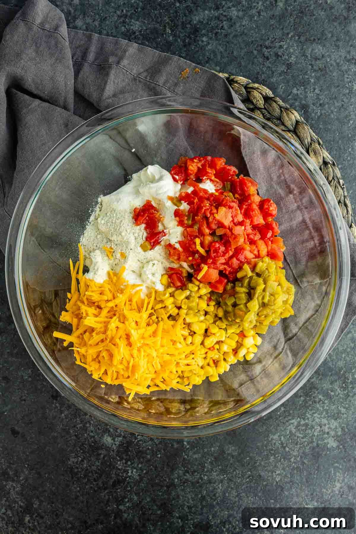 How to Make the Viral Boat Dip Everyone Loves 4 A glass bowl filled with shredded cheddar cheese, chopped green chilies, diced tomatoes, sour cream, and a dry seasoning mix sits on a dark surface with a gray cloth—an irresistible viral boat dip ready to be served.