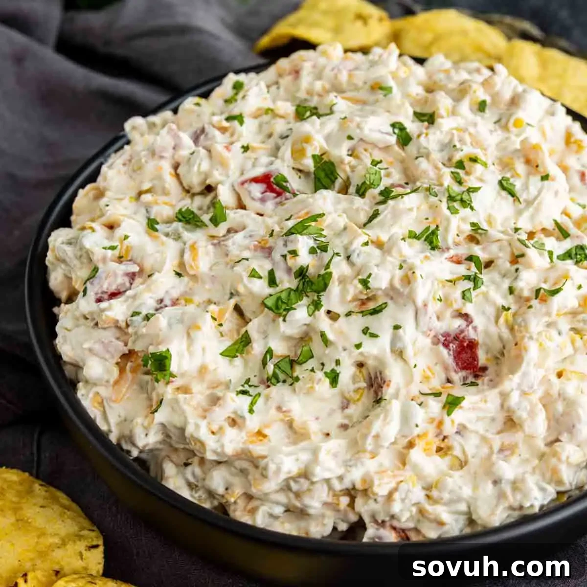 How to Make the Viral Boat Dip Everyone Loves 5 A black bowl filled with creamy cold corn dip containing chopped tomatoes and herbs, garnished with parsley, surrounded by tortilla chips.