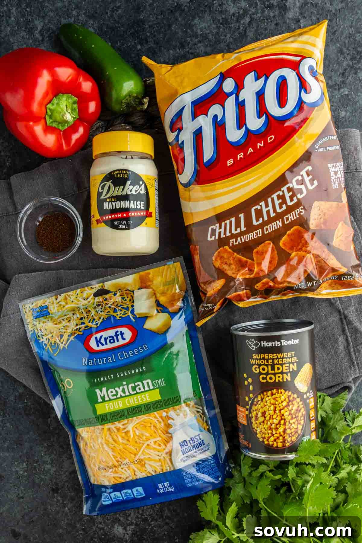 Frito Salad Recipe: Chili Cheese Corn Salad 3 A bag of chili cheese Fritos, shredded Mexican cheese, canned corn, mayonnaise, chili powder, red bell pepper, jalapeño, and fresh cilantro displayed on a dark surface—everything you need for an easy Frito salad recipe.