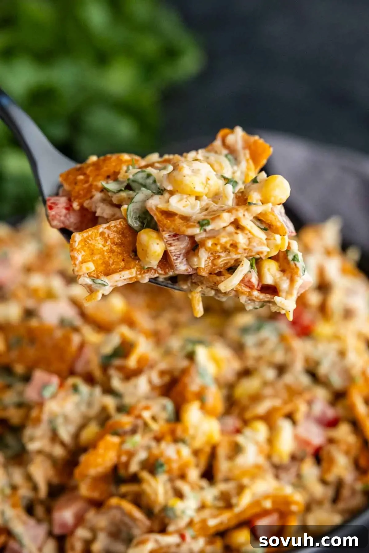 Frito Salad Recipe: Chili Cheese Corn Salad 5 A close-up of a fork holding a portion of corn chip salad with cheese, corn, diced tomatoes, herbs, and tortilla chips above a bowl of the same easy Frito salad recipe.