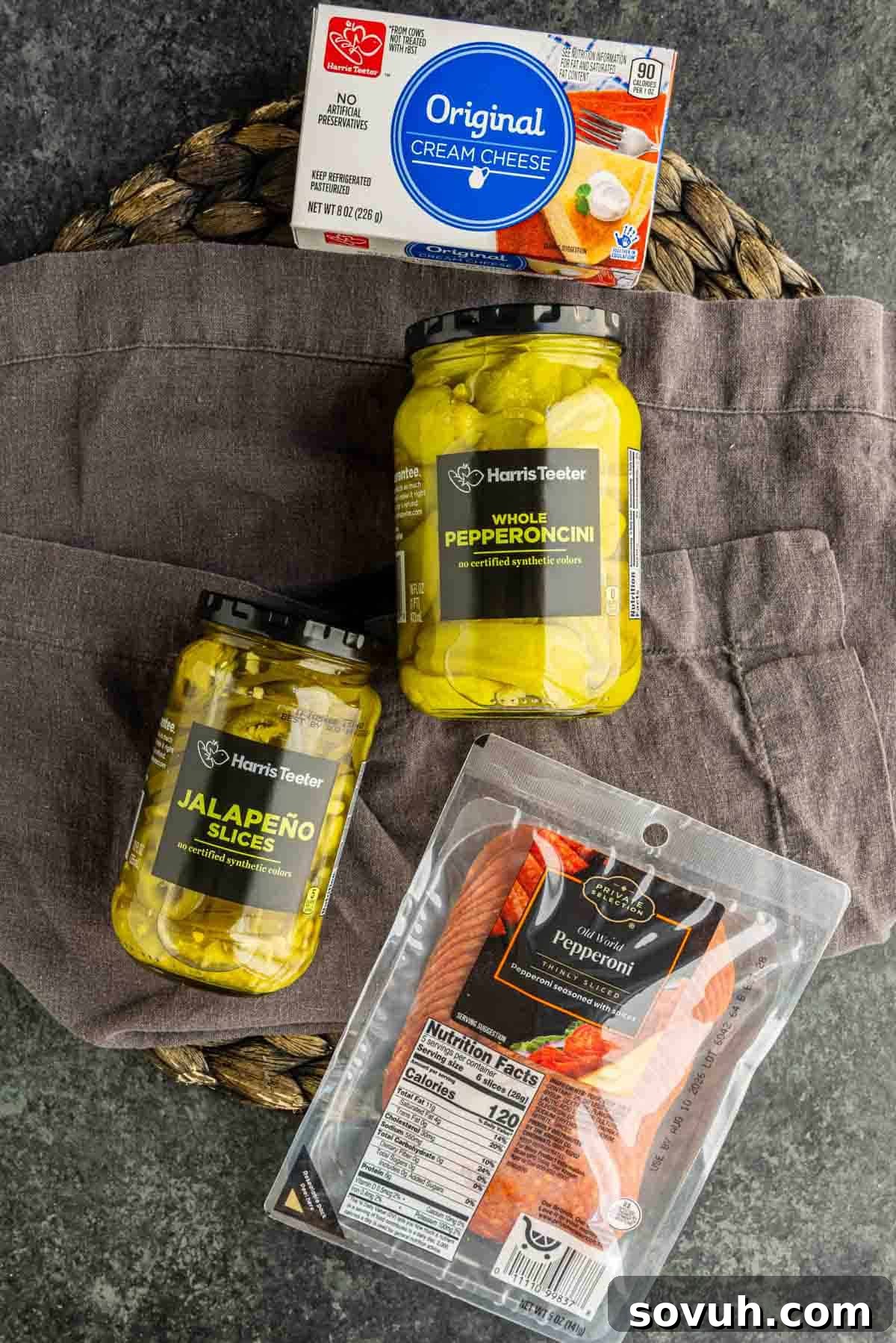 Air Fryer Stuffed Pepperoncinis with Pepperoni and Hot Honey 3 A package of cream cheese, two jars of pickled peppers, and a package of sliced pepperoni arranged on a gray cloth and woven mat—perfect ingredients for air fryer stuffed pepperoncinis.